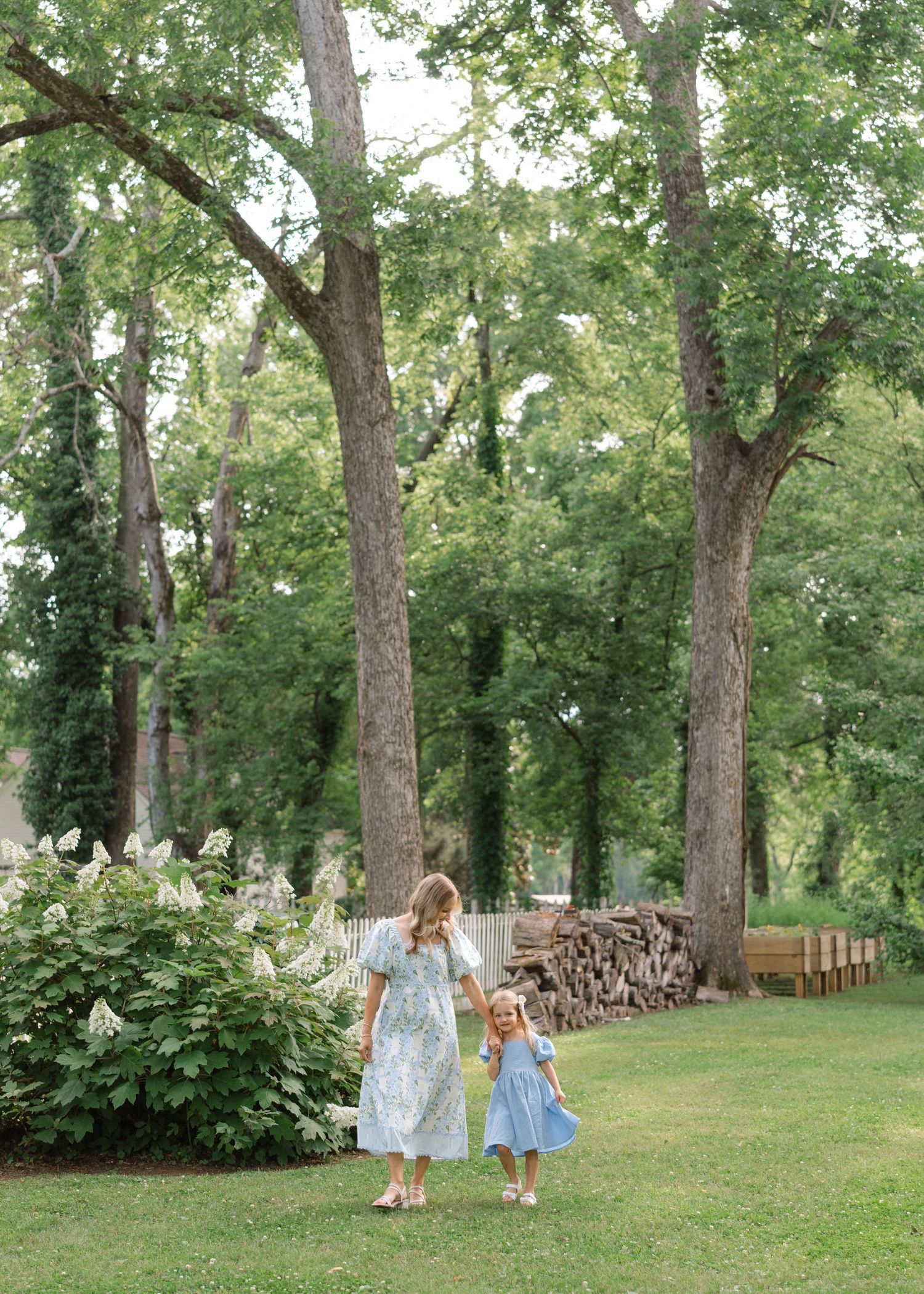 A mother and young daughter in matching blue dresses walk hand-in-hand through a lush green garden with tall trees.