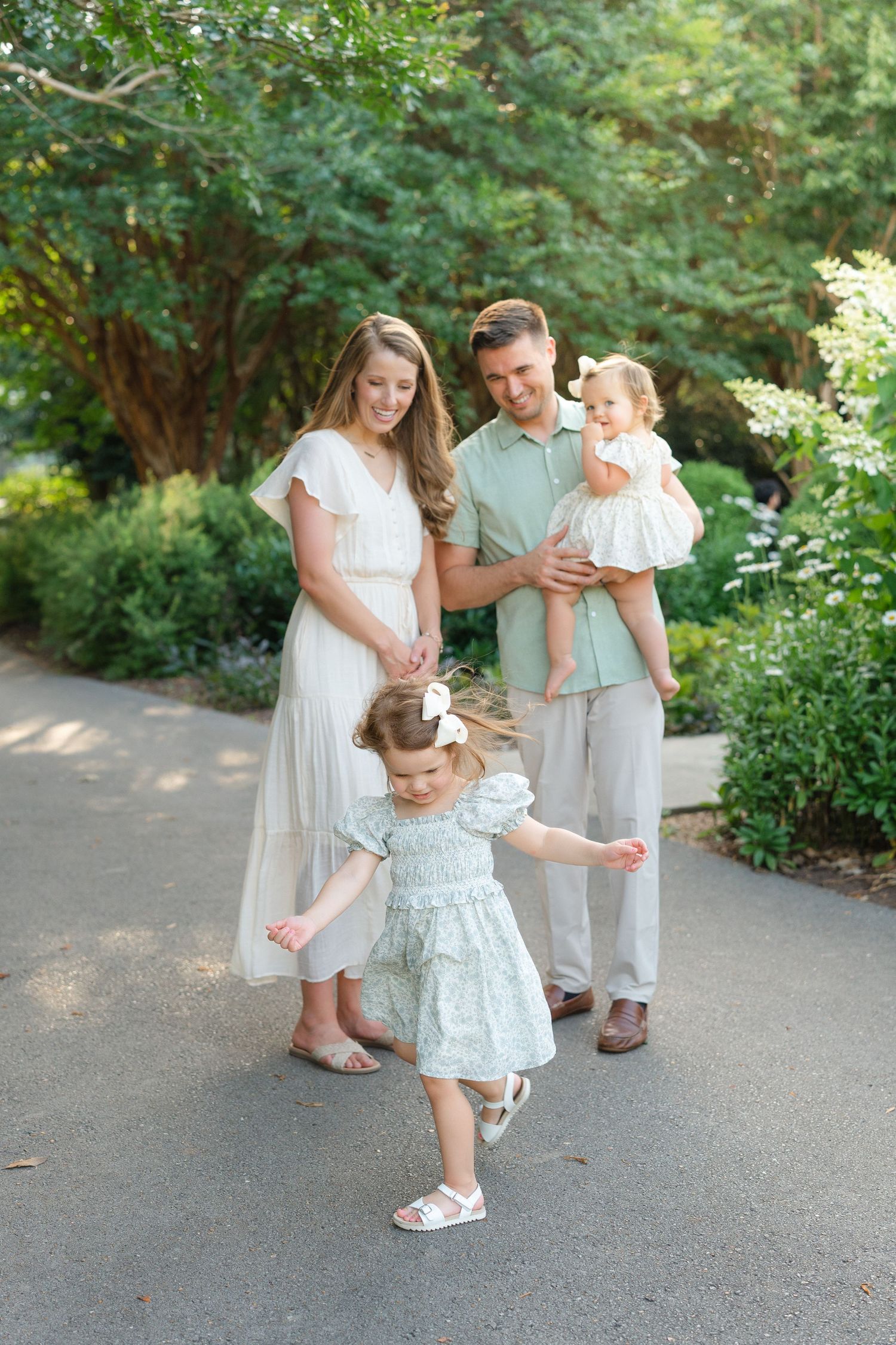 A happy family of four poses outdoors, with a pregnant mother, father holding a baby, and a toddler girl dancing on a garden path.
