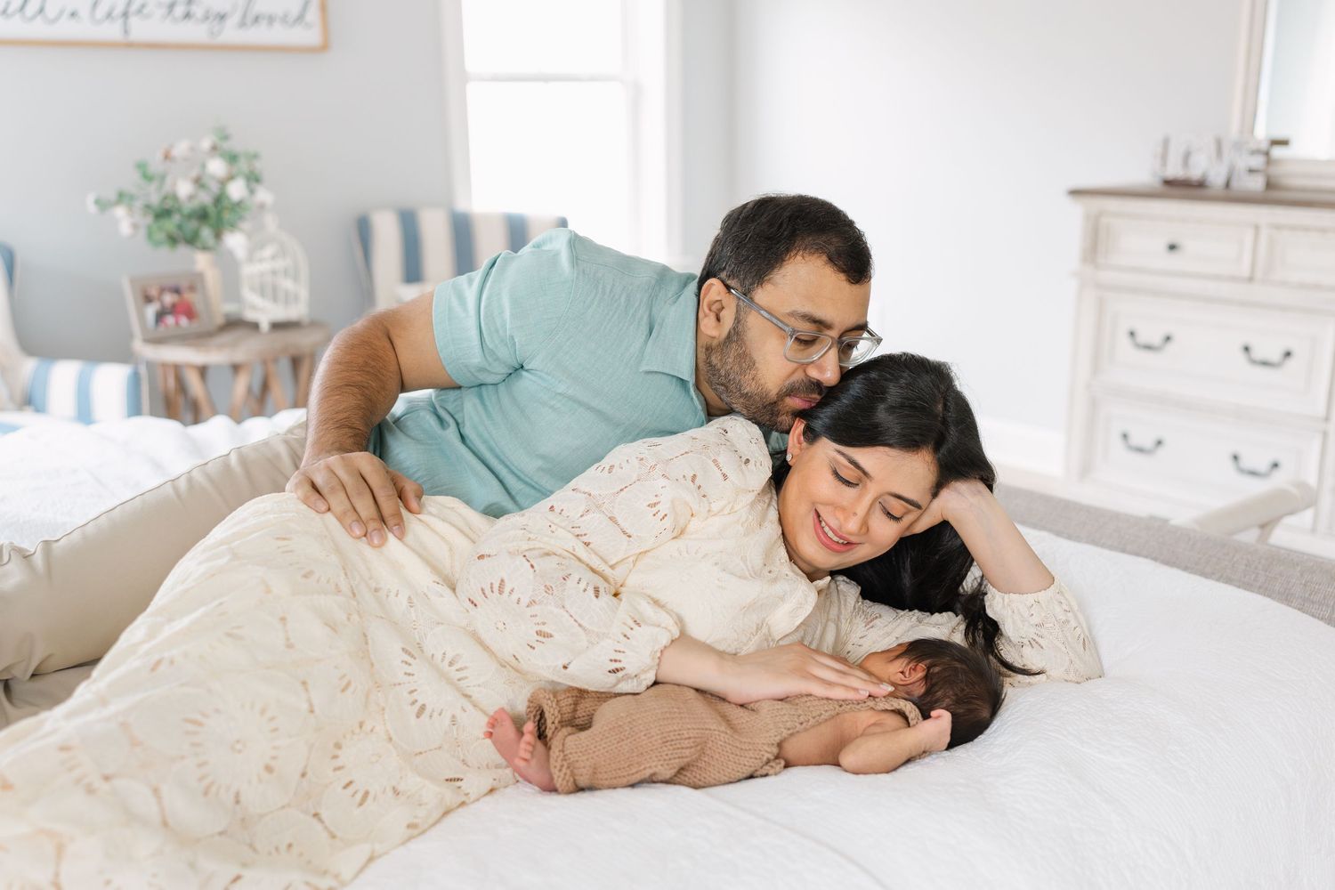 Two parents lean over their sleeping newborn baby lying on a white crocheted blanket on a bed.