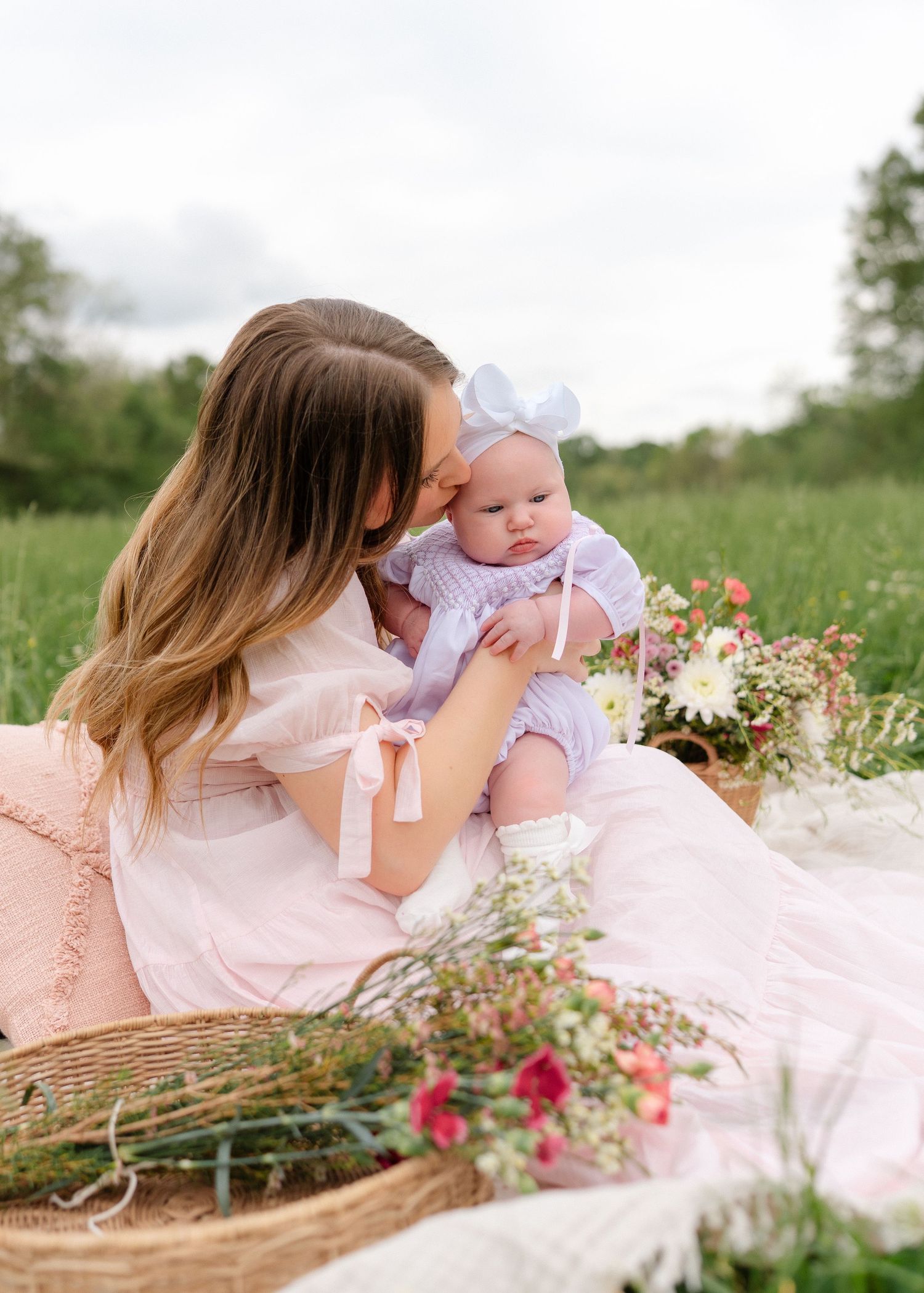 A mother in a pink dress kisses her baby girl wearing a white bow on a picnic blanket outdoors.