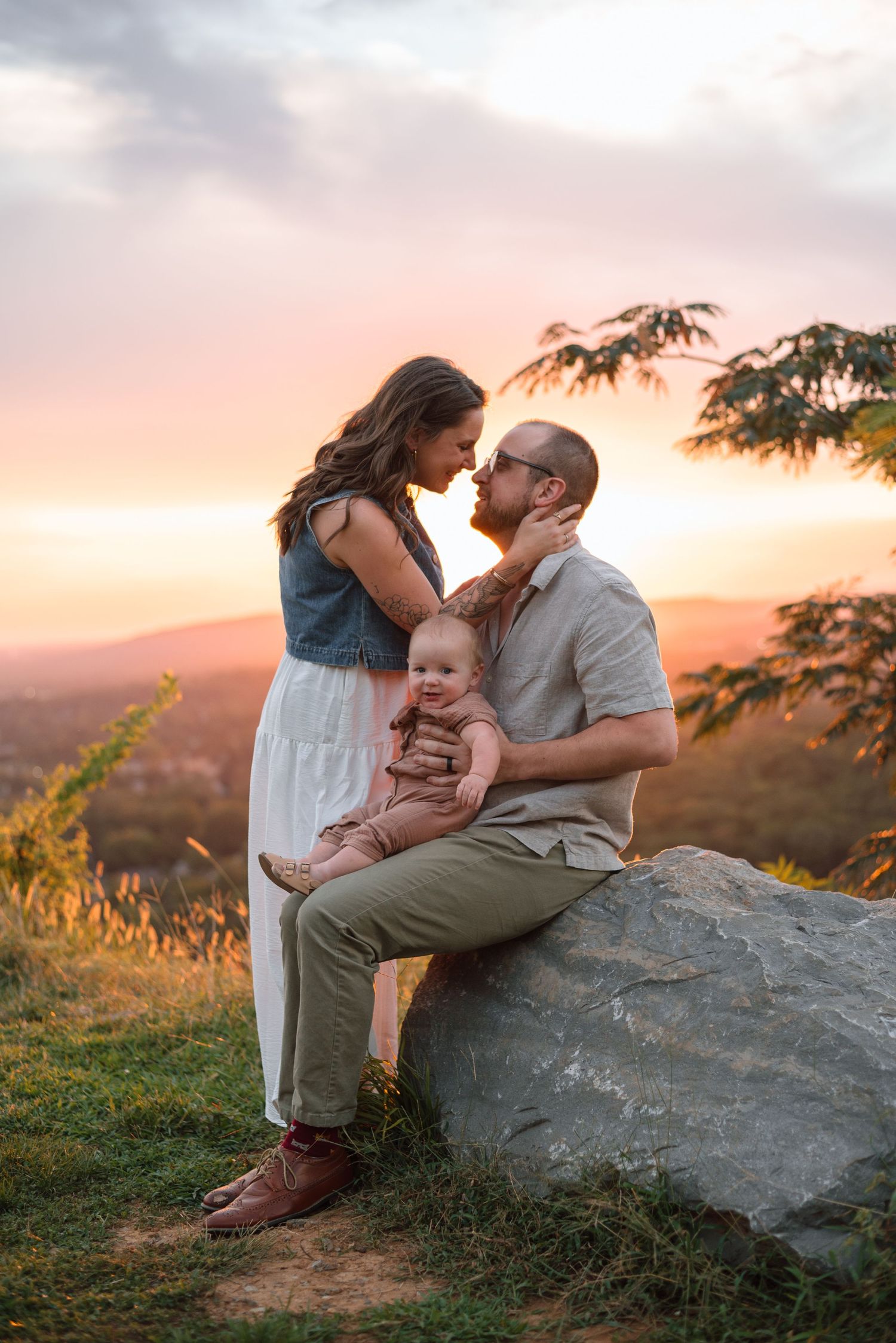 A couple leans in for a kiss while their baby sits on a rock during a colorful sunset.