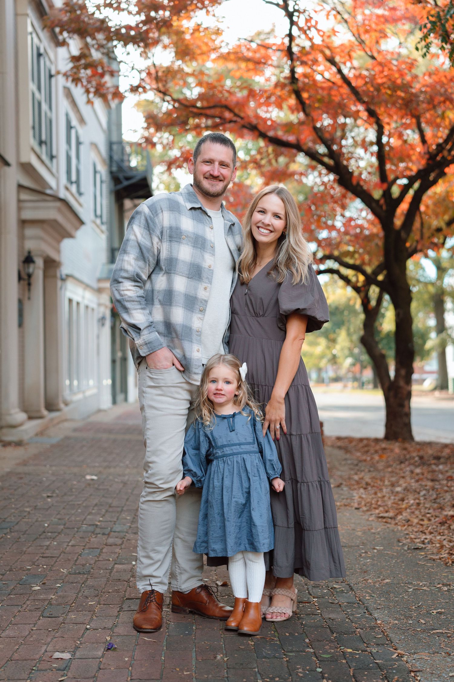 A smiling family of three poses on a brick sidewalk surrounded by vibrant red fall foliage trees.