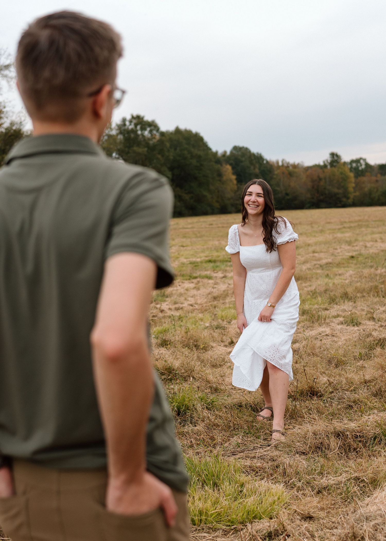 Smiling woman in white dress standing in an open field facing her partner outdoors.