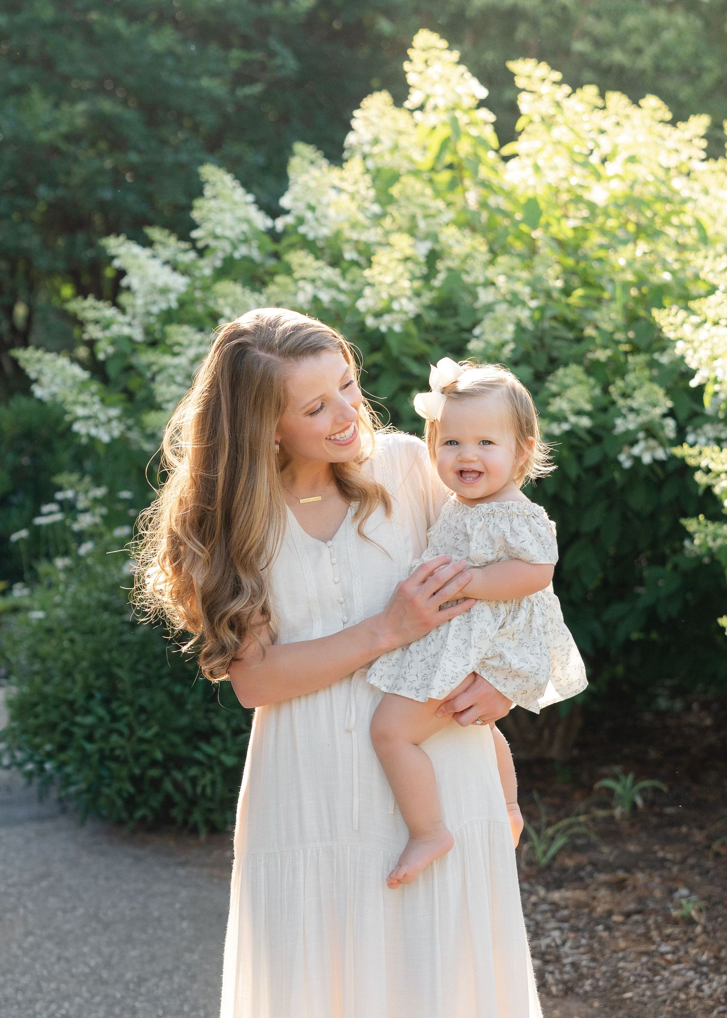 A smiling mother in a white dress holds her happy toddler girl outdoors in a sunlit garden.
