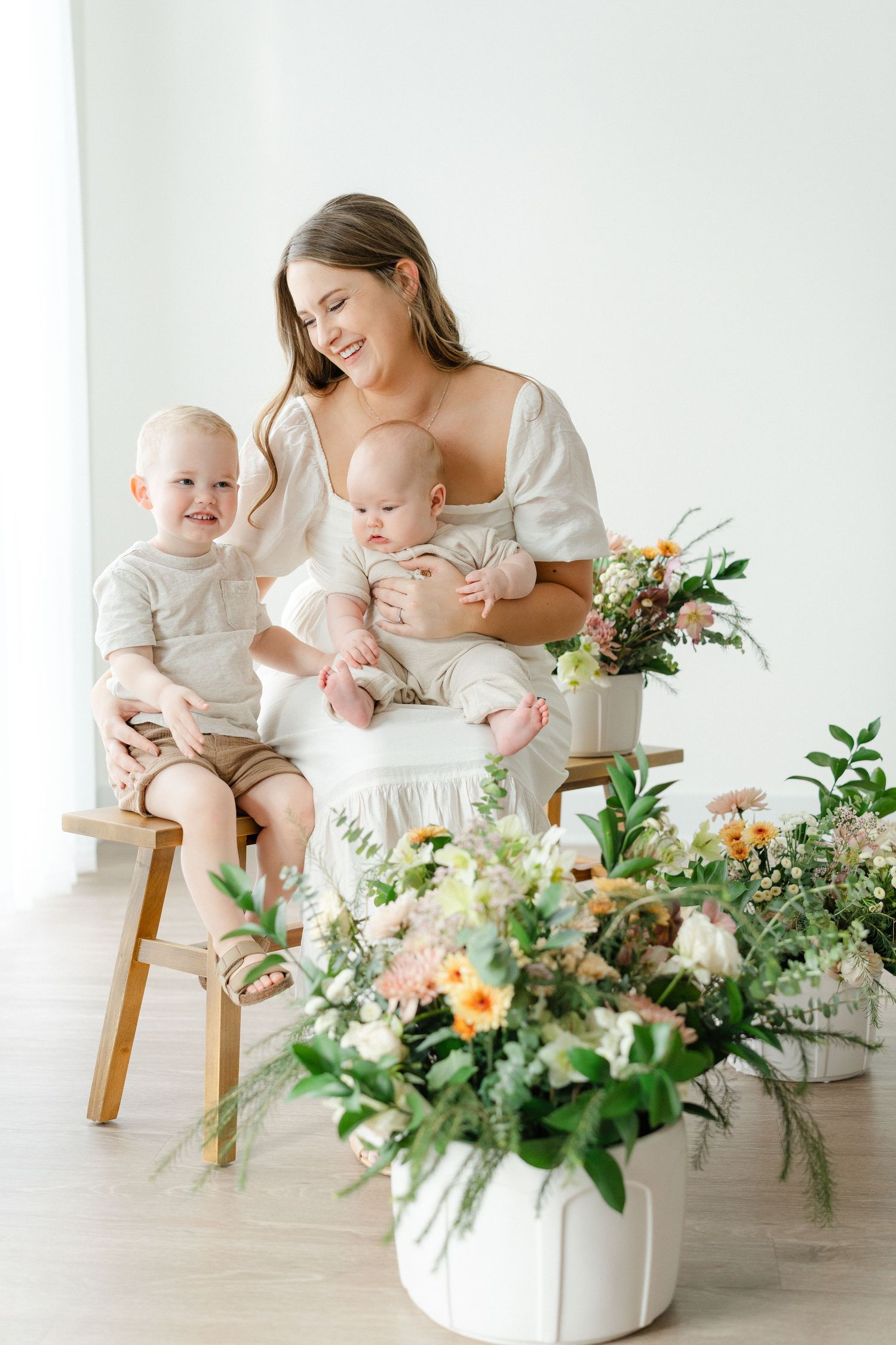 Happy mother in white dress sits on wooden bench holding two young children with flowers nearby.