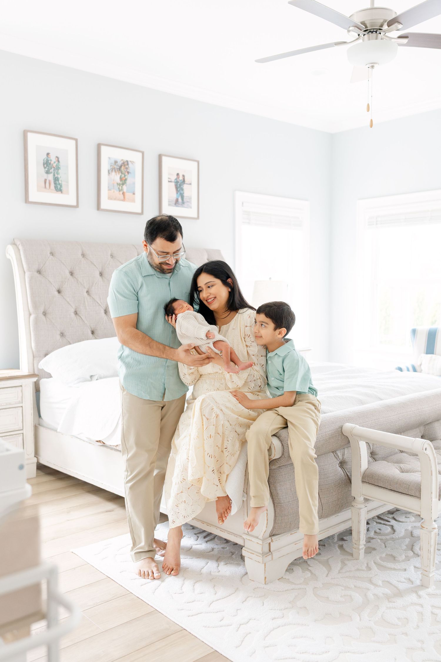 Family of four with newborn poses together on bed in bright bedroom with framed photos.