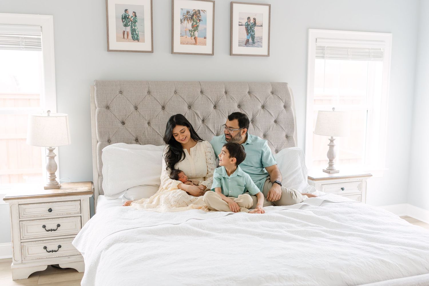 A happy family with a toddler and newborn sits together on a white bed in a bright bedroom.
