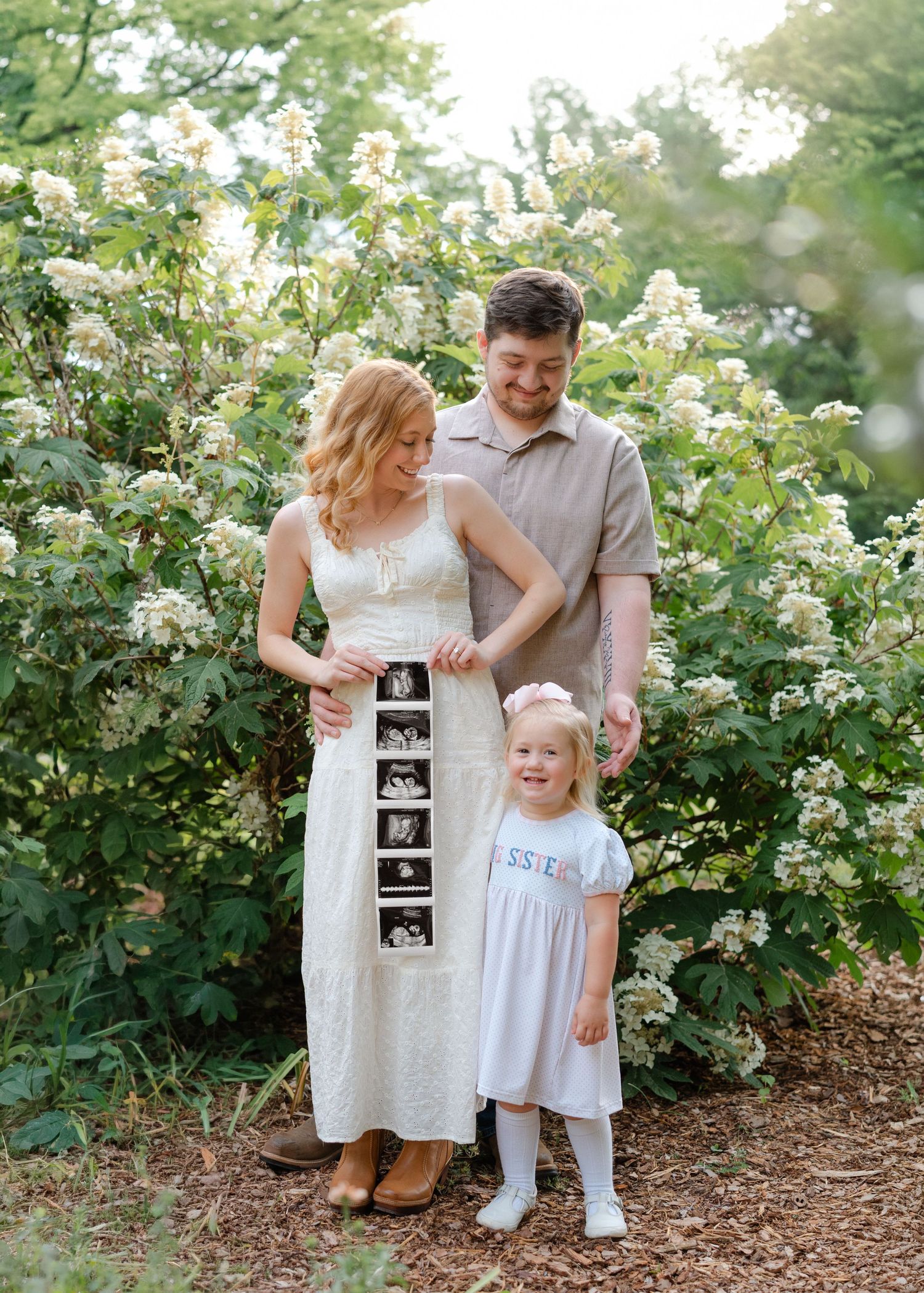 Pregnant couple holding ultrasound photos with toddler girl wearing Sister shirt standing in front of white flowering bushes.