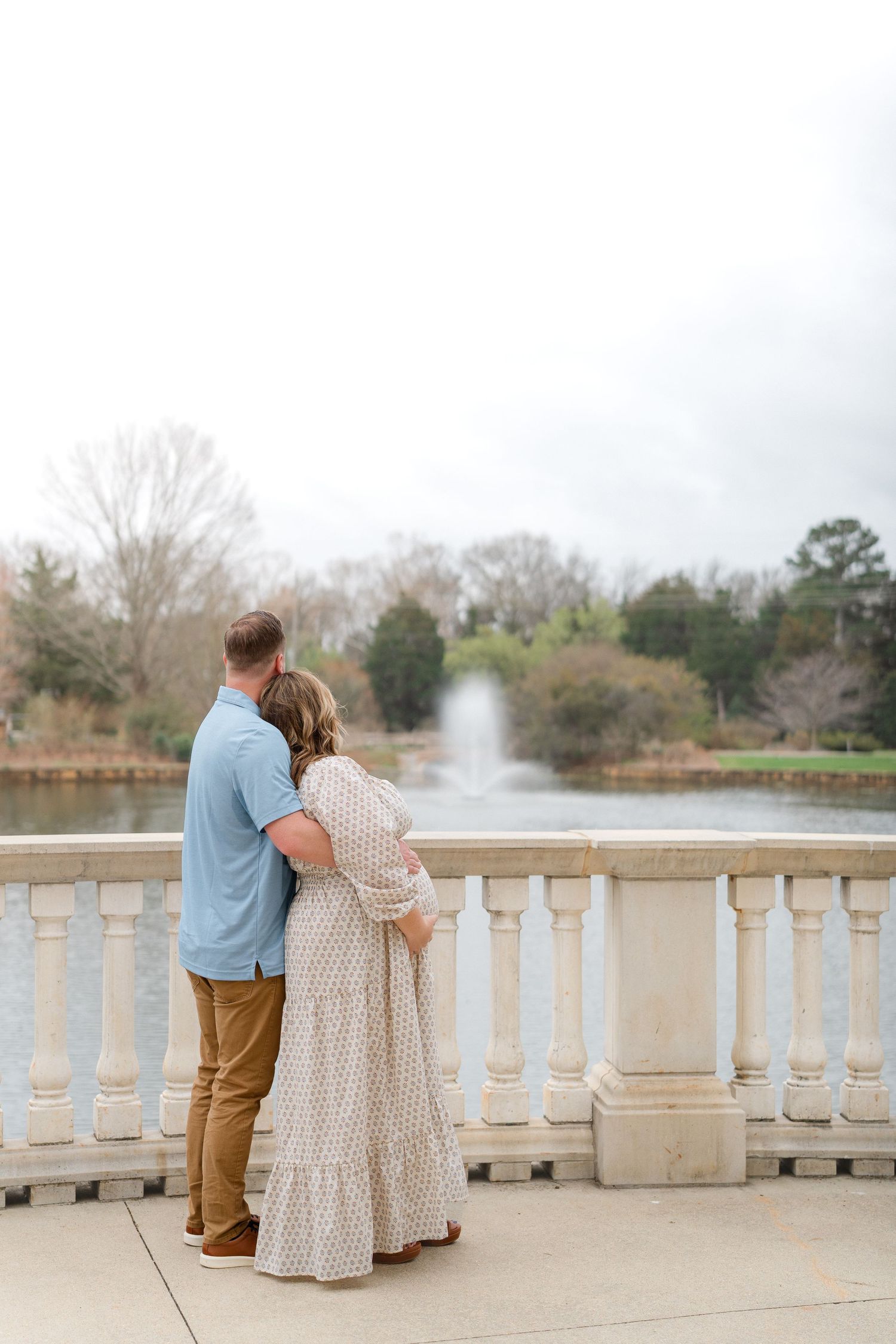 A pregnant couple stands embracing on a balcony overlooking a serene pond with a fountain in a park.