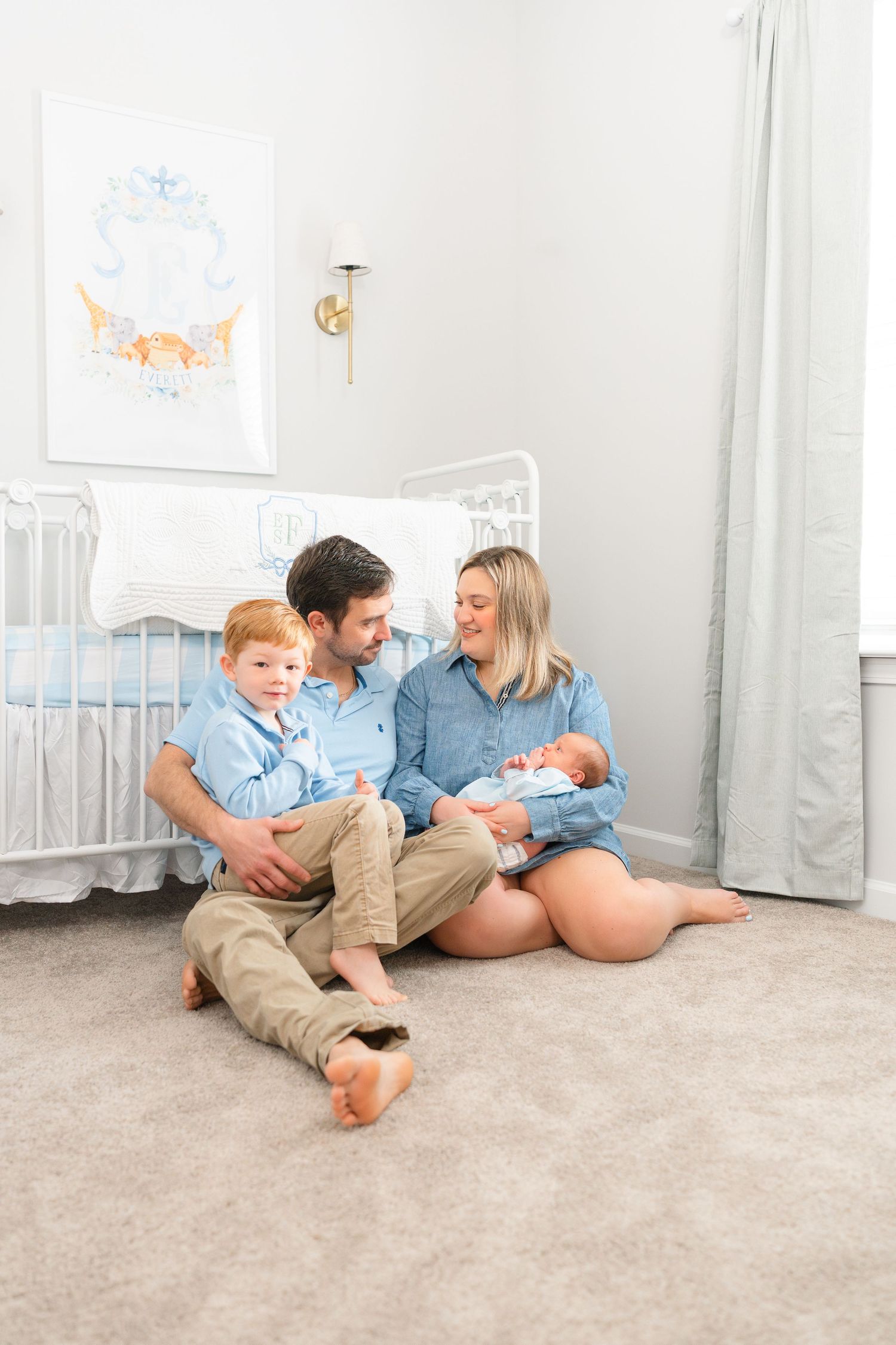 Family of four sitting on nursery floor with newborn baby and red-haired toddler son.