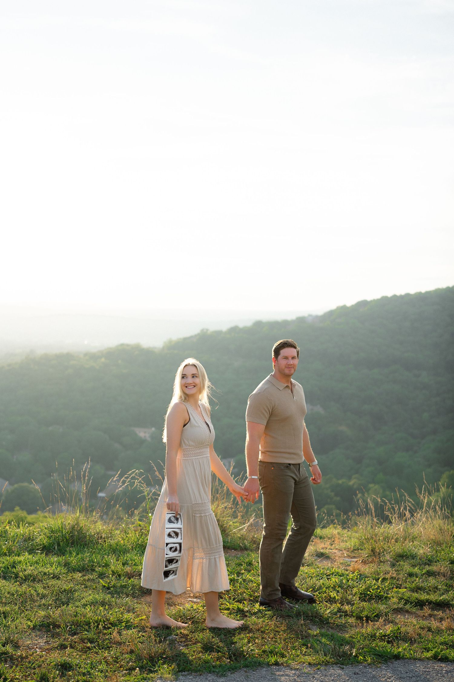 Couple holding hands on a grassy hilltop at sunset, woman in white dress, man in khaki, with misty green mountains behind.