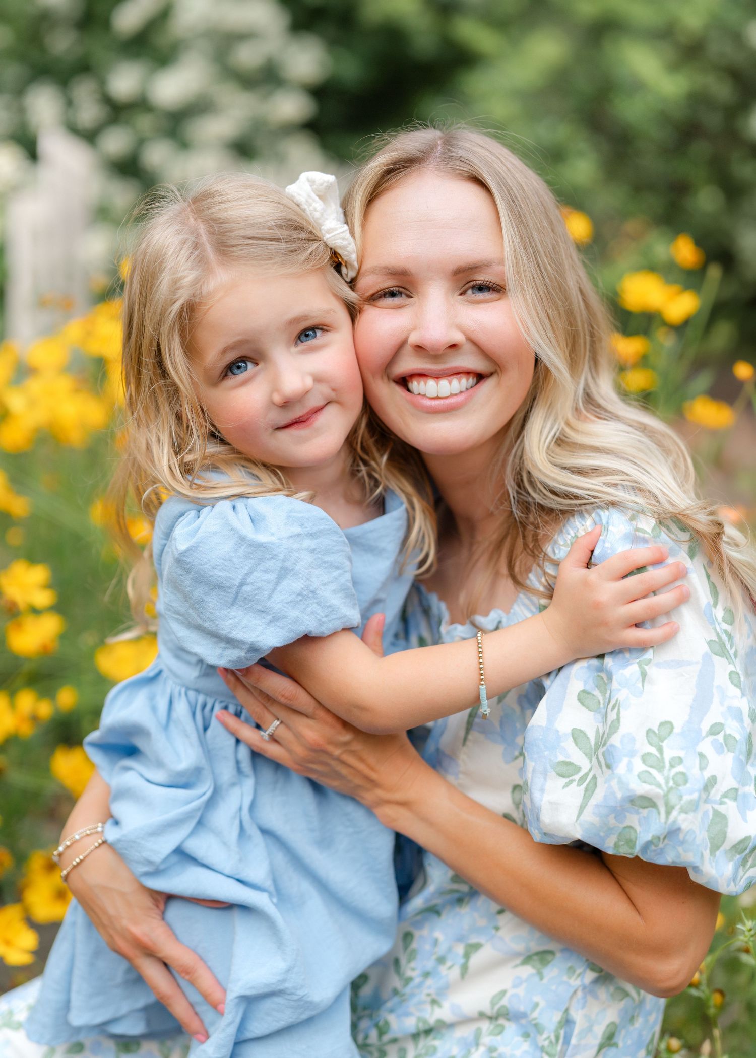 A smiling woman hugs a young blonde girl in a blue dress among yellow wildflowers in a sunny garden.