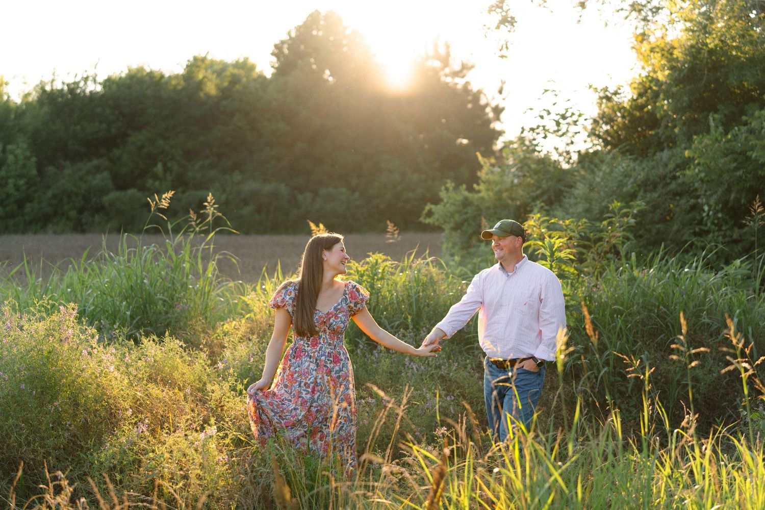 Couple holding hands walking through a golden sunlit meadow, woman in floral dress and man in white shirt and jeans.