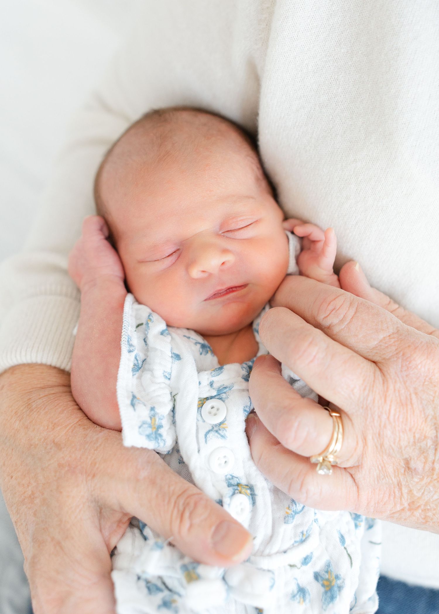 Sleeping newborn baby in floral onesie cradled gently in grandparent's hands with gold ring visible.
