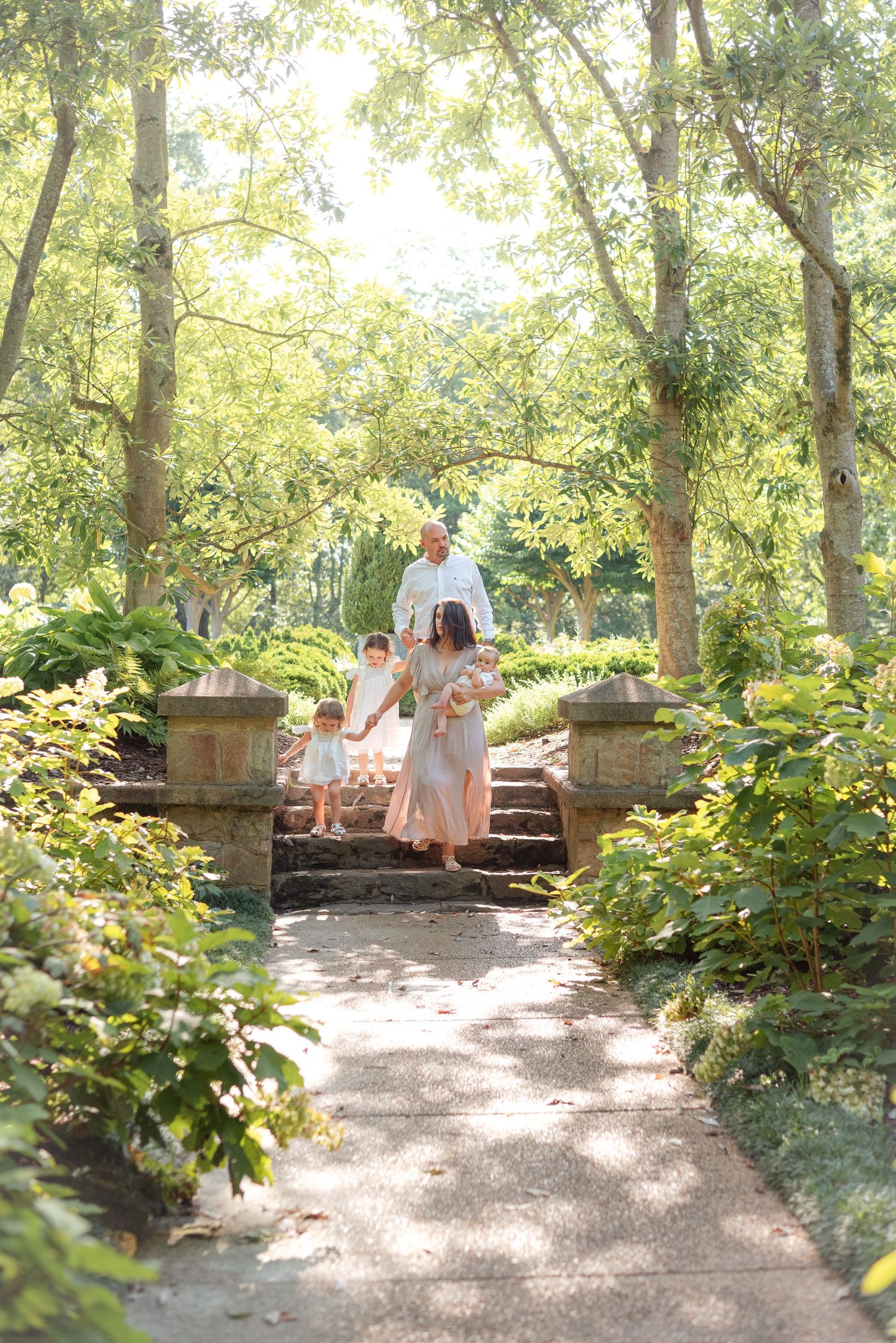 Family of four walking down sunlit stone garden steps surrounded by lush green trees and ferns.