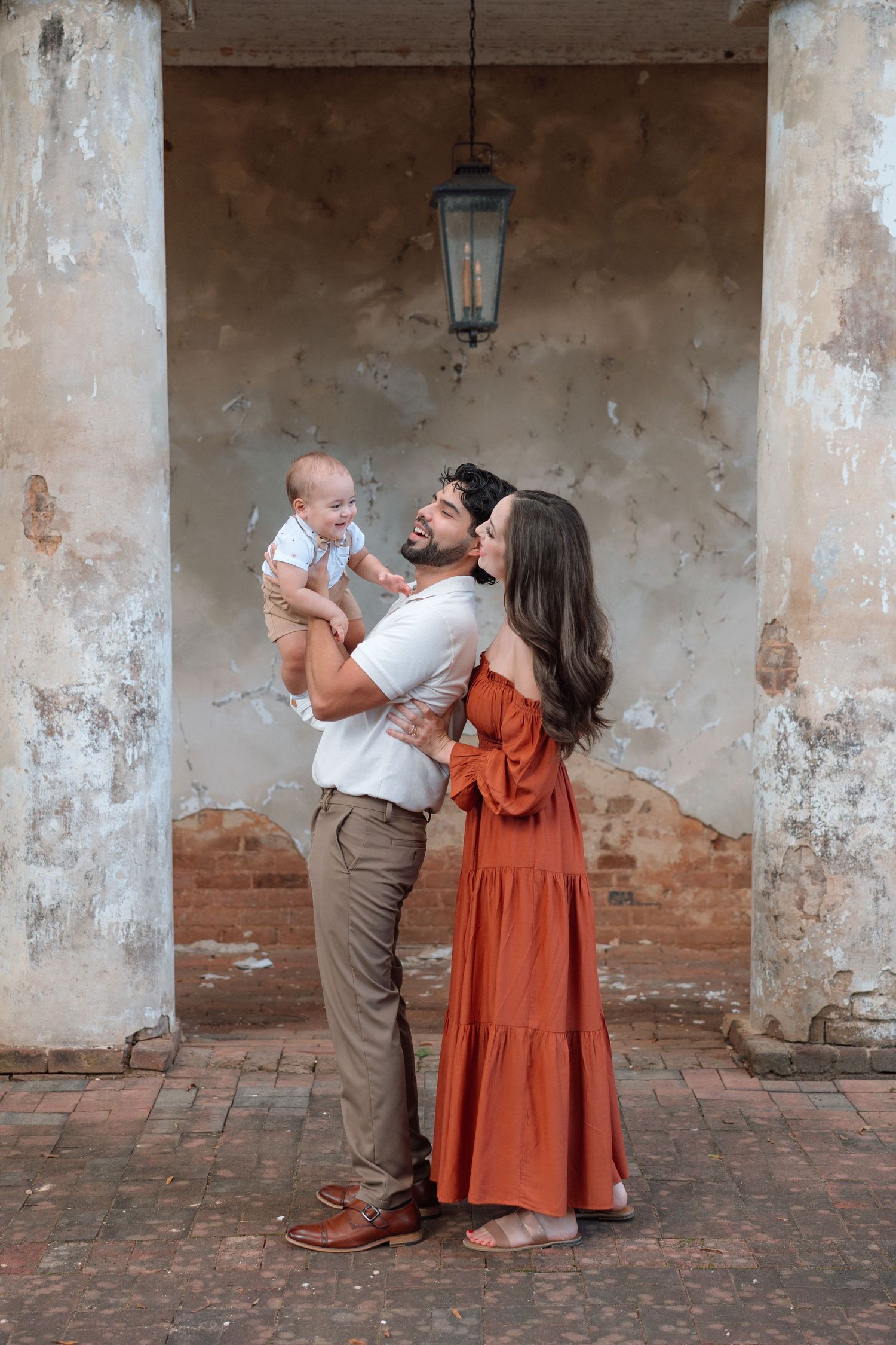 A couple holding their baby shares a tender moment under a rustic archway with brick walls.