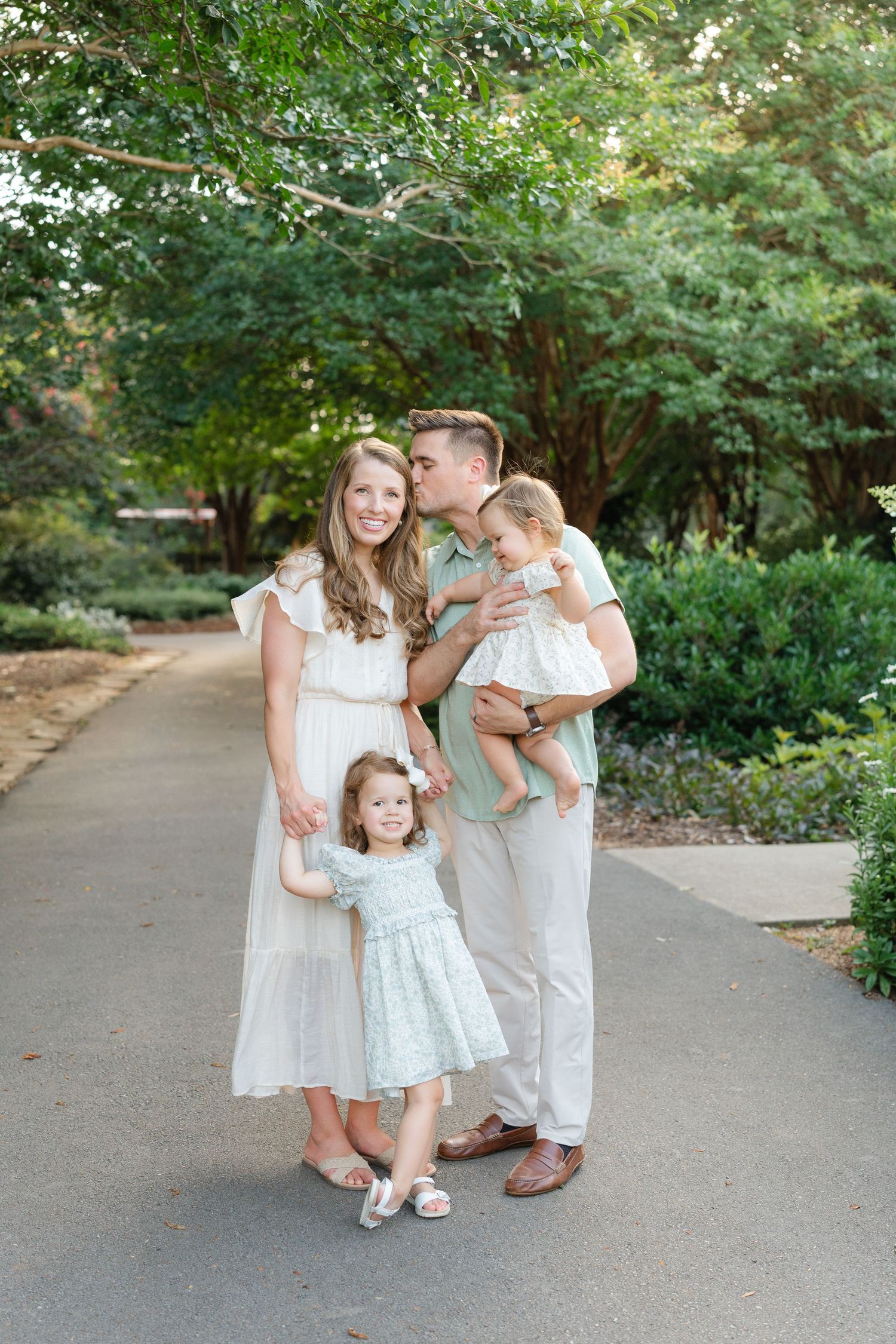 Family of four posing outdoors, mother in white dress, father kissing baby, toddler girl in blue dress.
