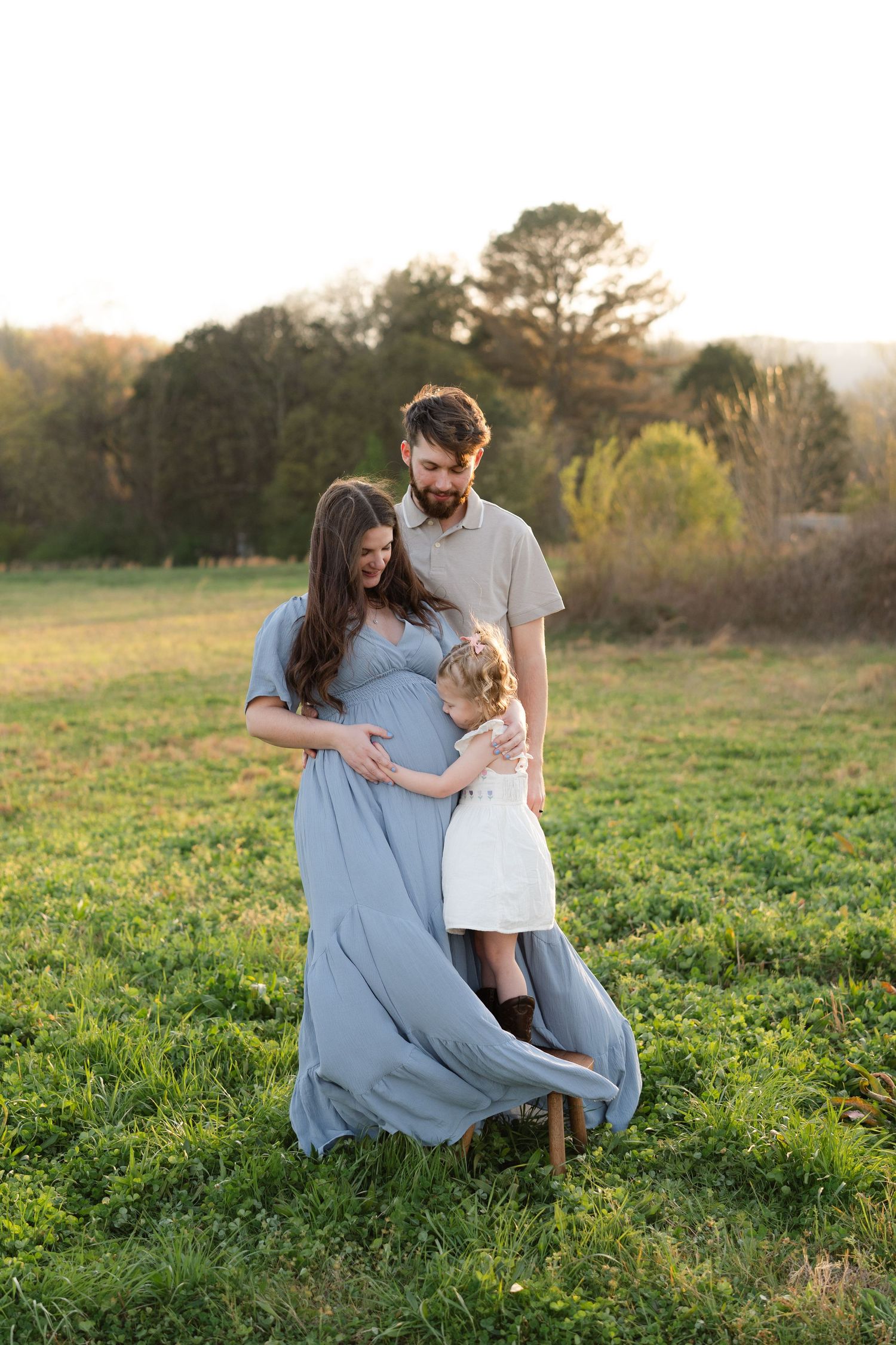 A pregnant woman in a blue gown stands with her partner and young daughter in a lush green field at sunset.