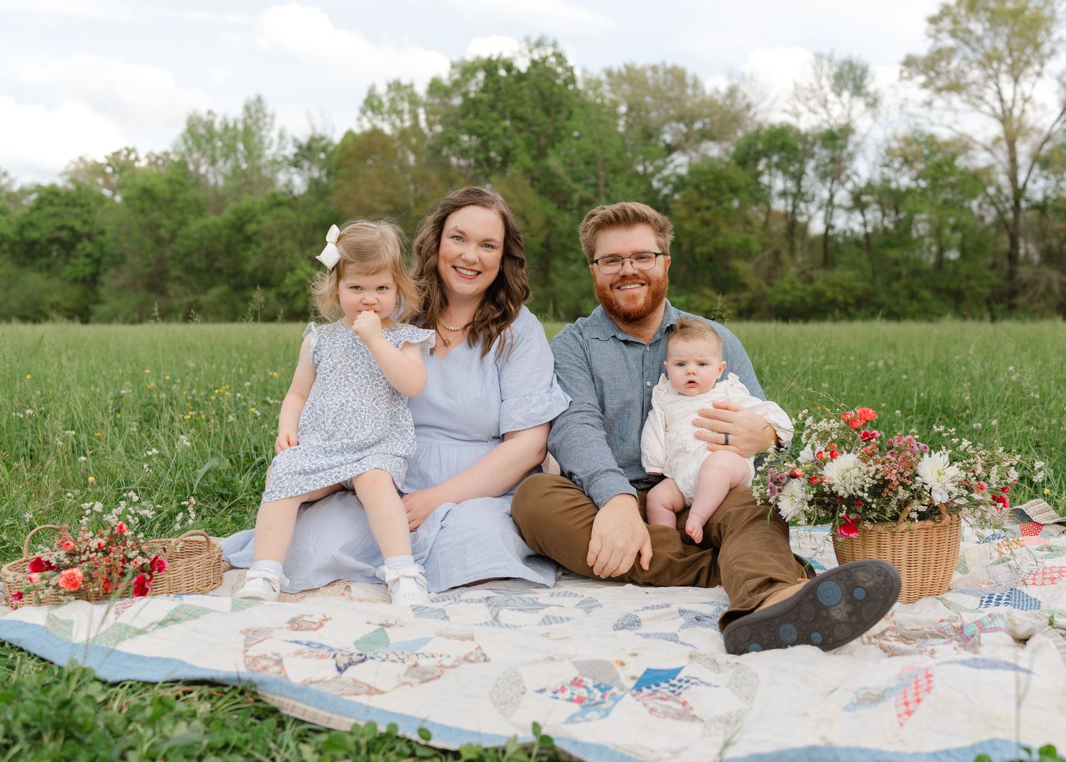 A happy family of four sits on a quilt in a green field surrounded by wildflowers and a wicker basket.