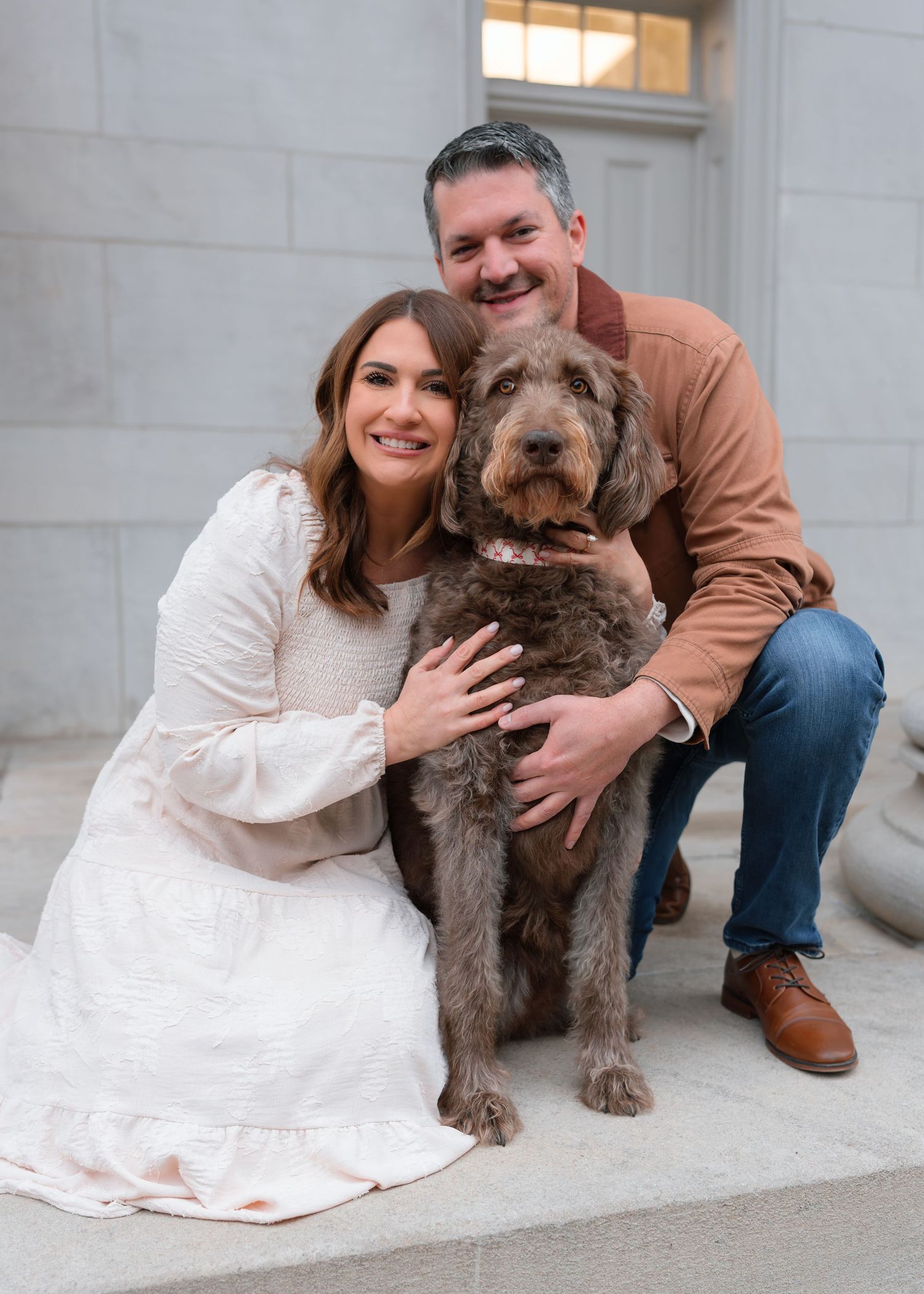 Smiling couple poses with their chocolate labradoodle on stone steps near a classical building.