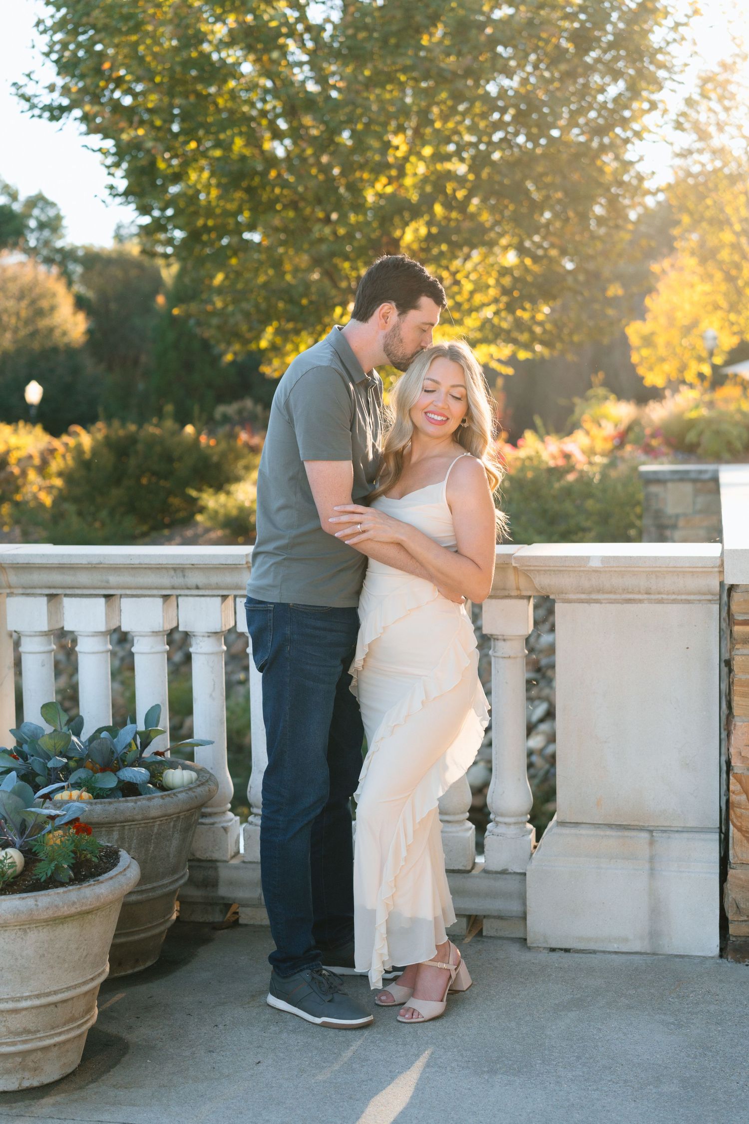 Couple embracing on elegant stone balcony at golden hour, woman in cream dress smiling joyfully.