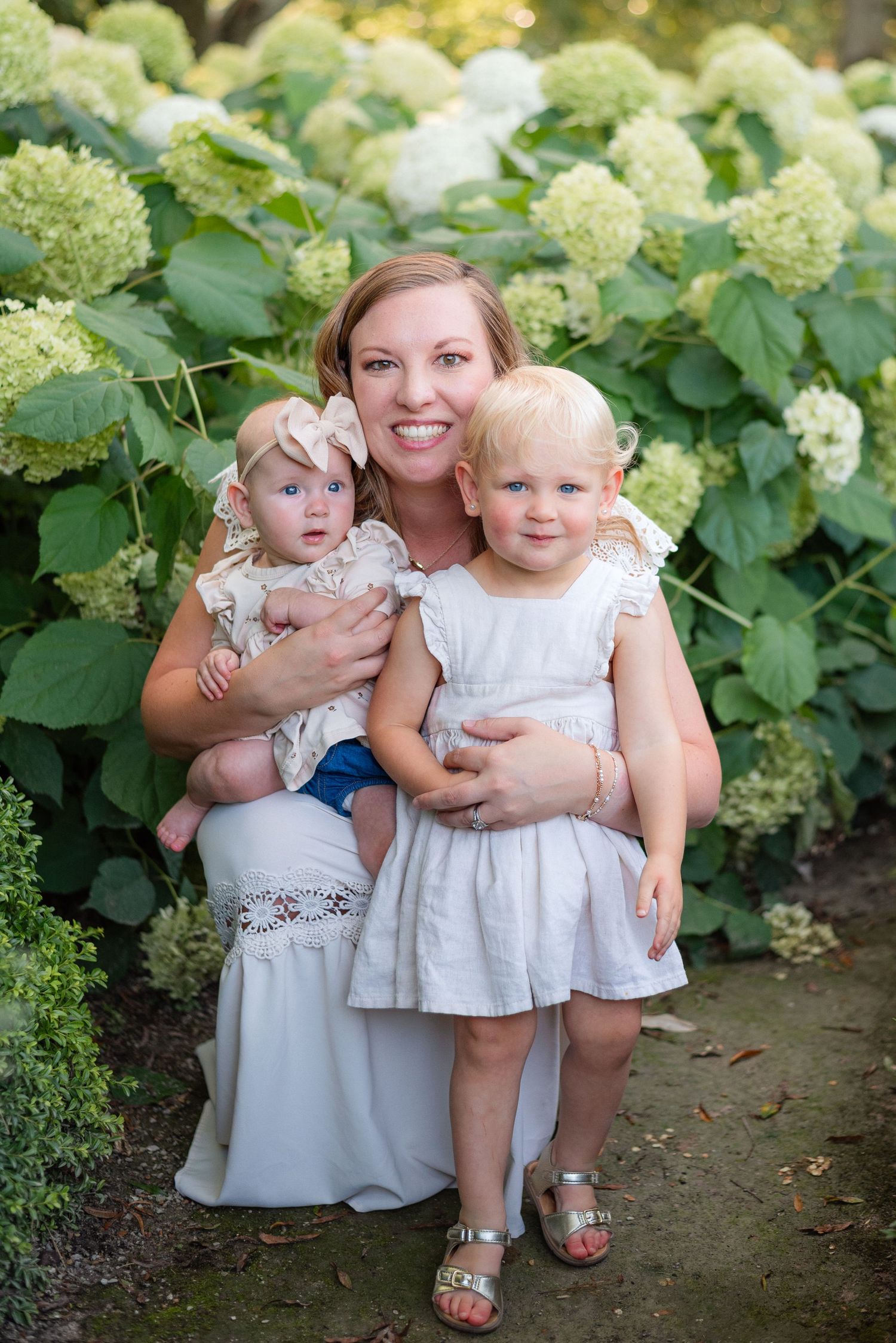 Smiling mother kneels with two young daughters in white dresses before lush white hydrangea bushes.