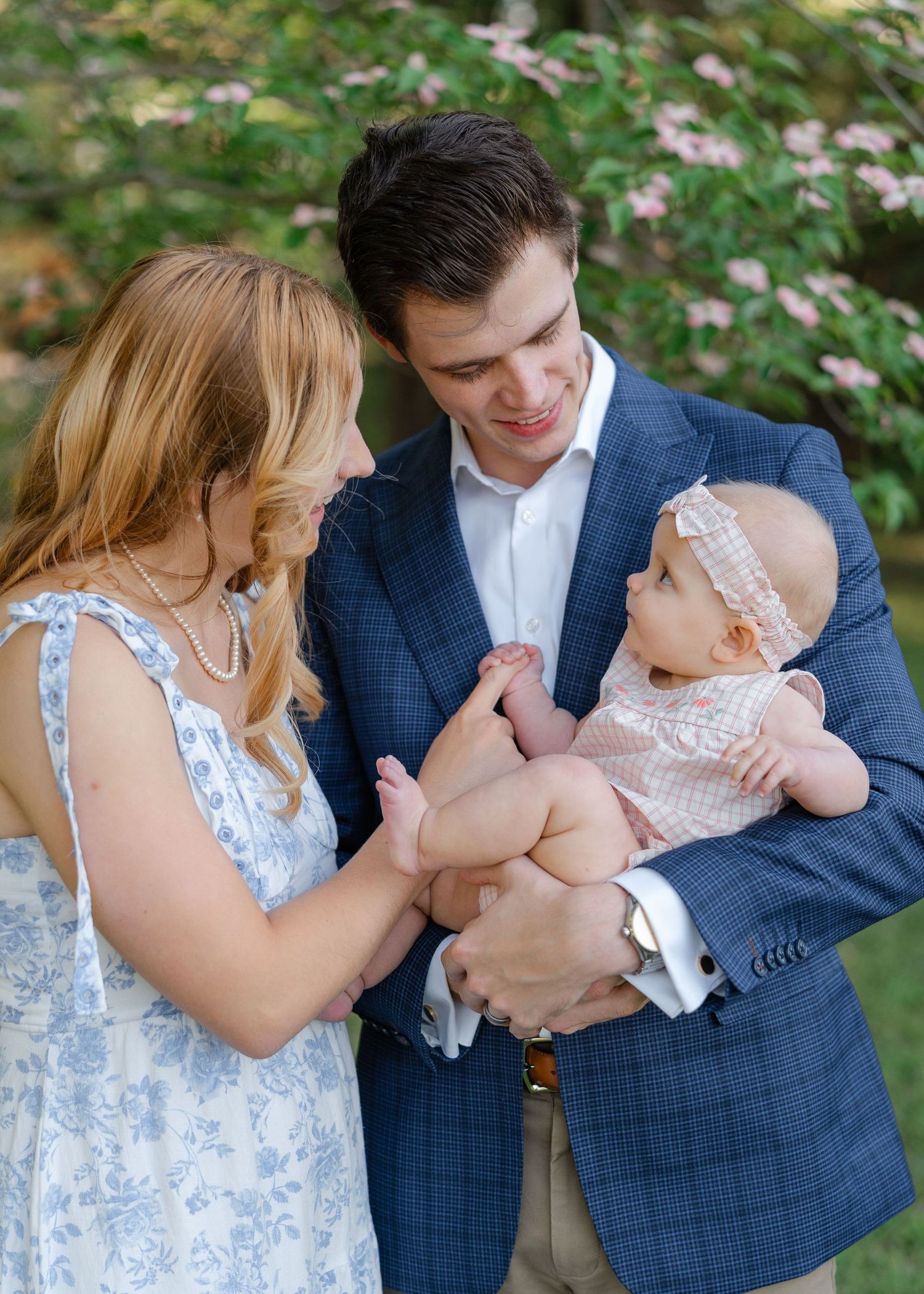 Young couple in elegant attire smiles at their baby girl during an outdoor photo session.