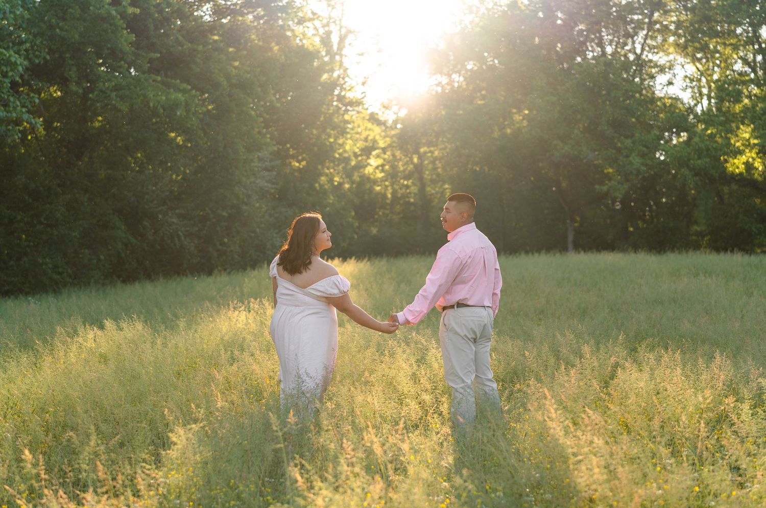 Couple holding hands in a sunlit meadow surrounded by lush green trees during golden hour.
