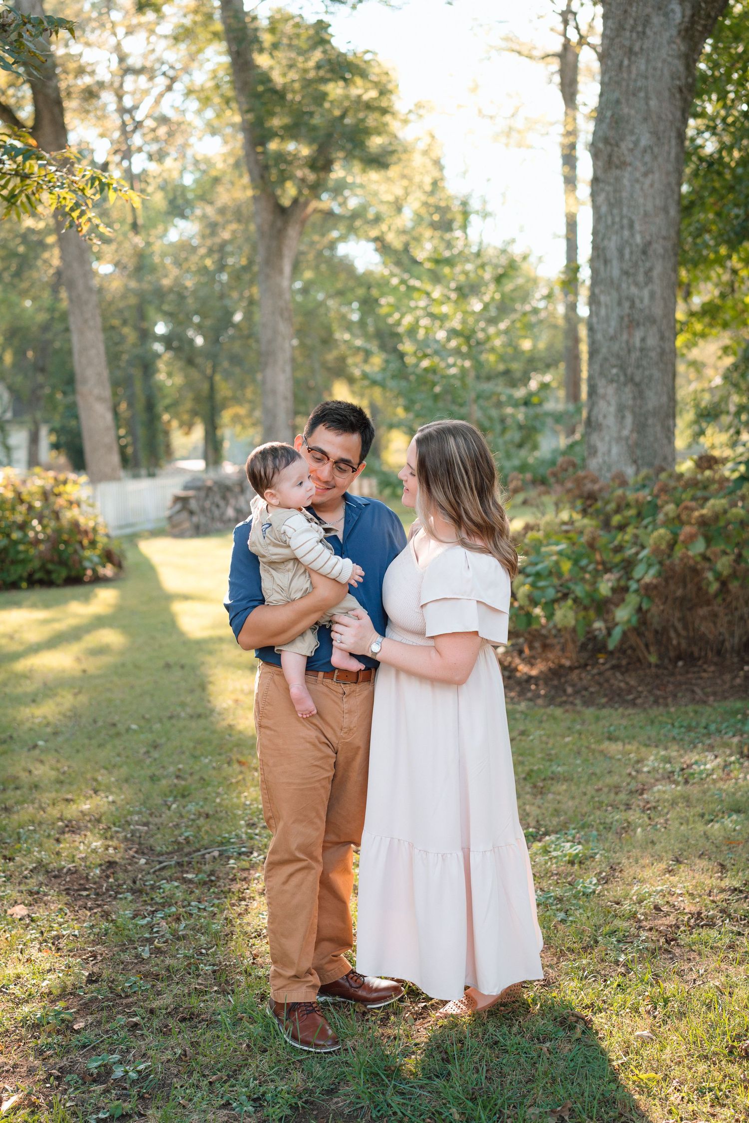 A couple stands outdoors among tall trees, the man gently holding their baby while facing his wife.