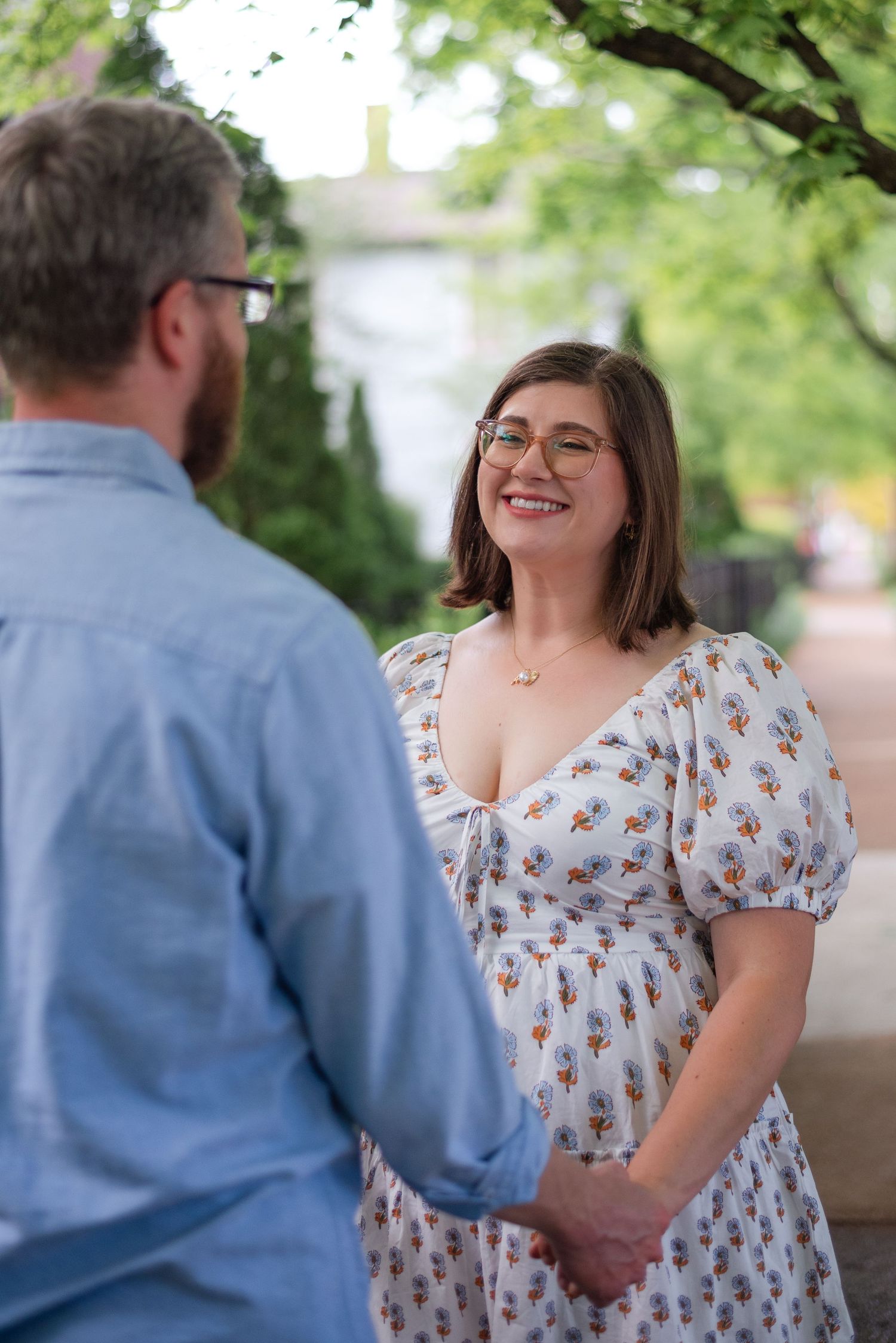 A smiling woman in a floral dress holds hands with a man during an outdoor engagement session.