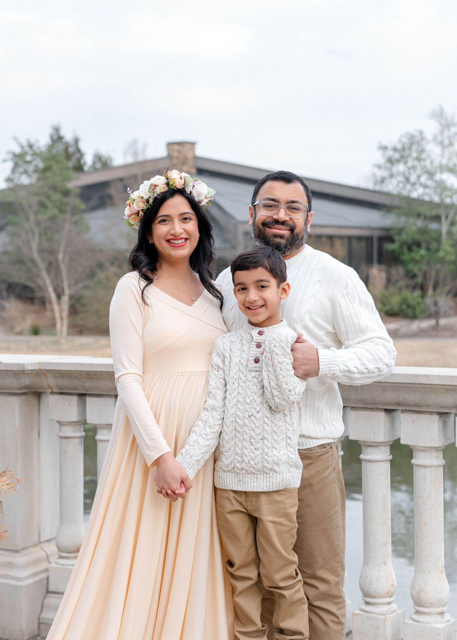 Pregnant woman in cream gown with floral crown poses with husband and son outdoors.