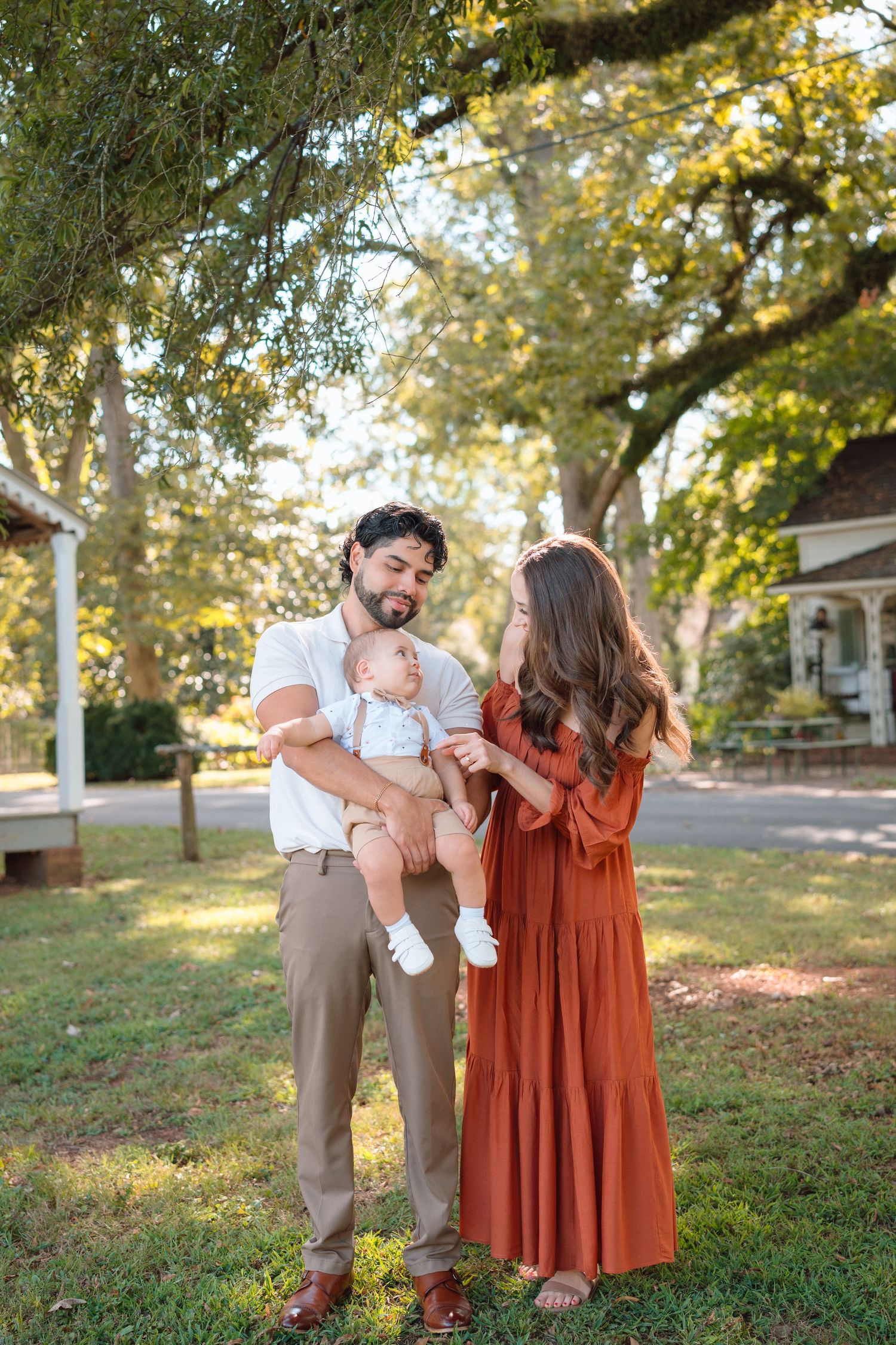 A young family with a baby boy poses outdoors under large oak trees, mom in rust dress, dad in white shirt.