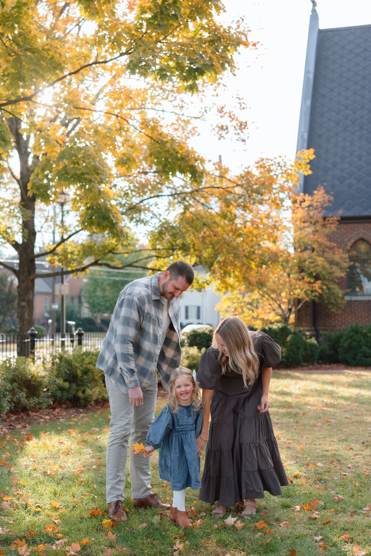 A family of three enjoys autumn foliage, parents smiling at their young daughter in a park.