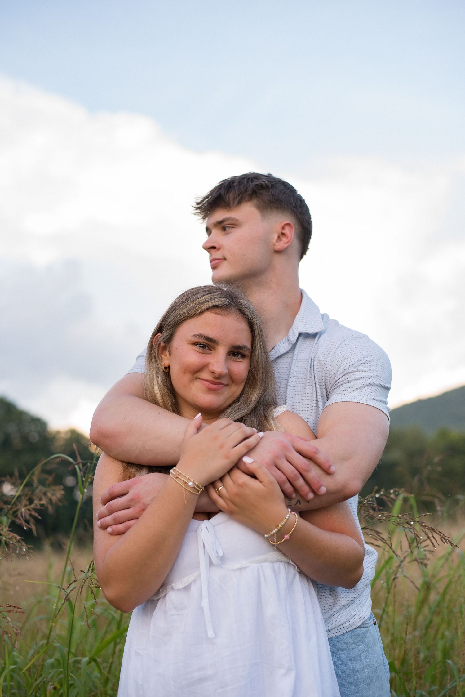 Man hugging woman from behind in a golden field with mountains and cloudy sky backdrop.