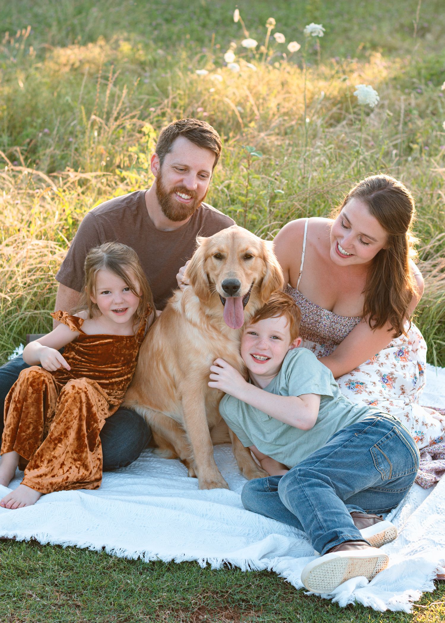 Happy family with two children and a golden retriever sitting together on a blanket in a sunny meadow.