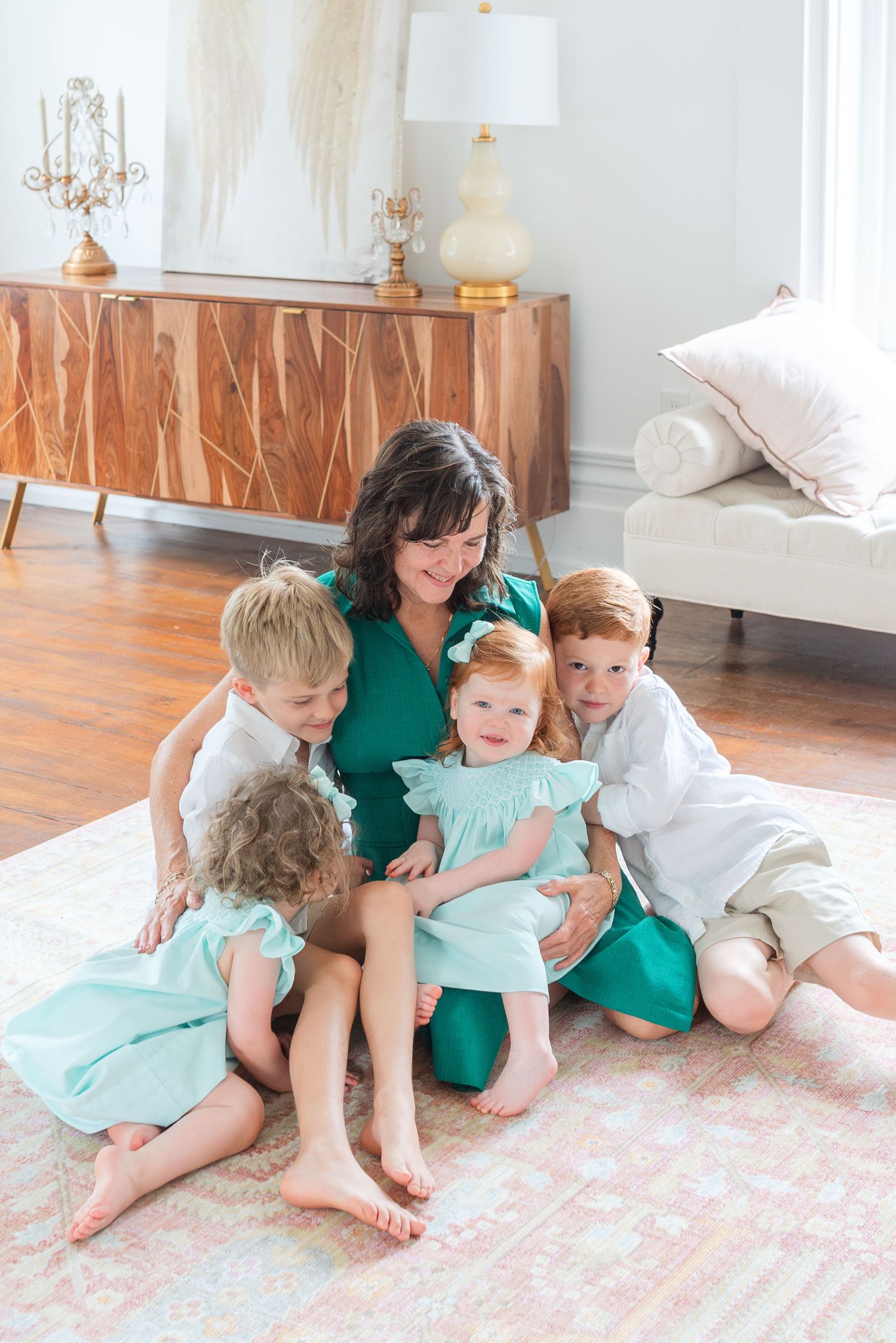Mother in green dress surrounded by four young children in mint outfits on a pink rug indoors.