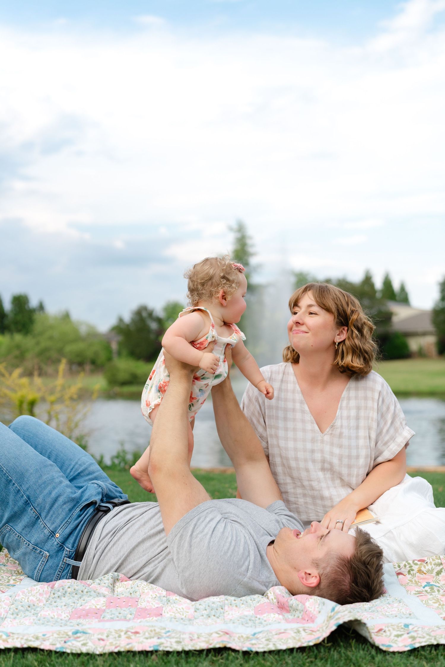 Happy family enjoying a picnic on a quilt, parents playing with baby outdoors near a pond.