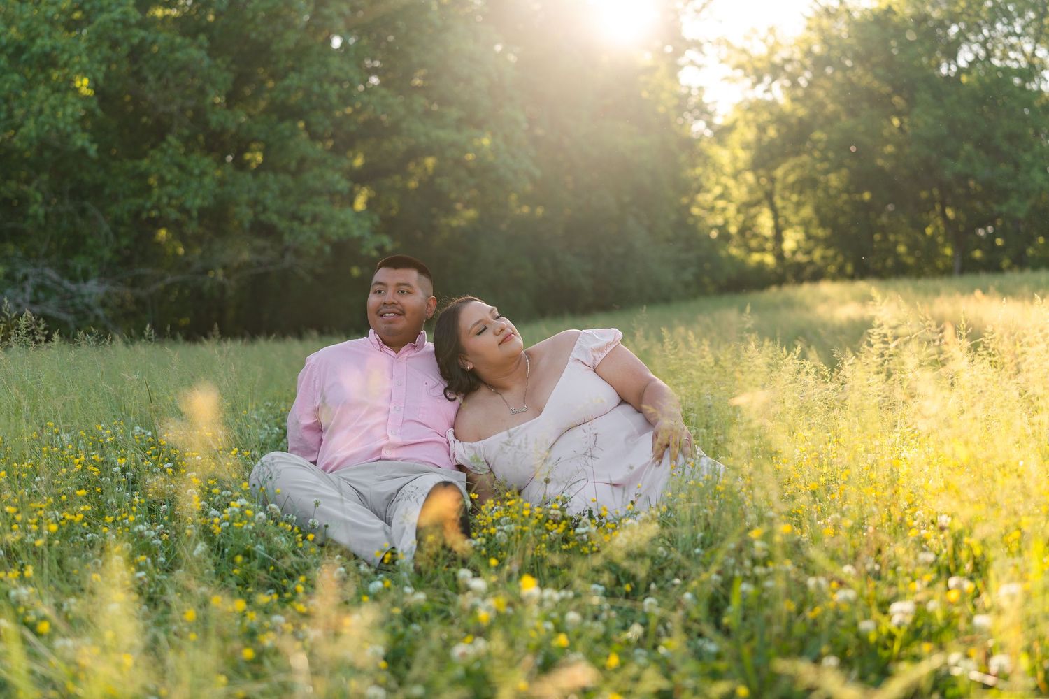 Couple sitting in a sunny meadow with wildflowers, laughing together in golden hour light.