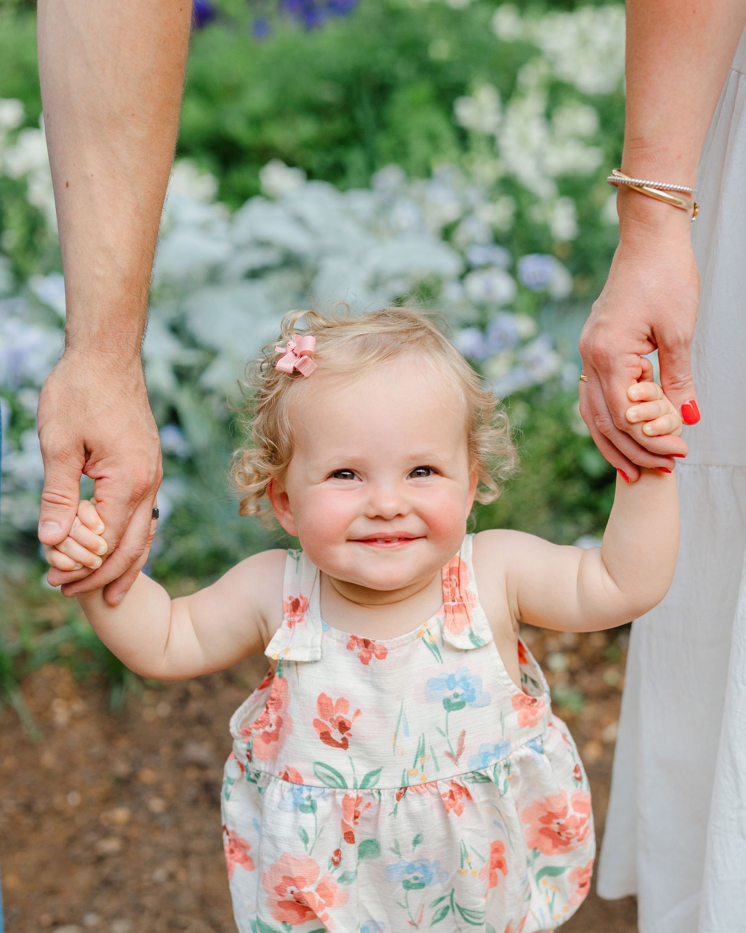 Smiling toddler girl in floral dress holds parents hands outdoors among blooming garden flowers.