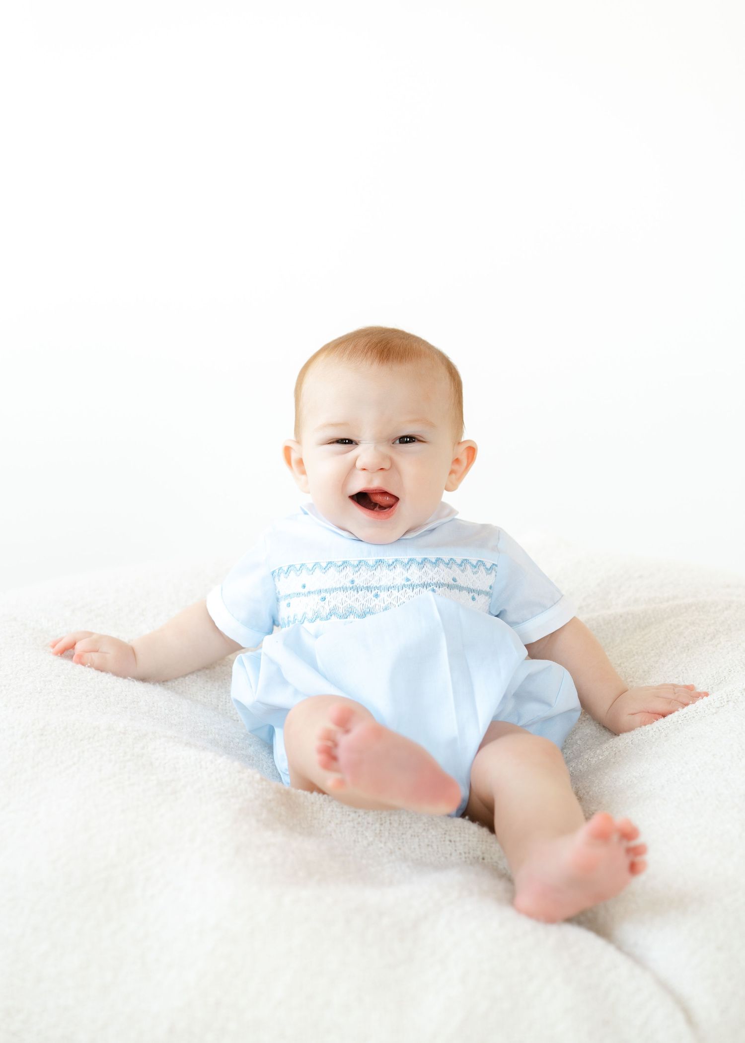 Smiling baby boy sitting upright wearing a light blue smocked romper on a white fluffy blanket.