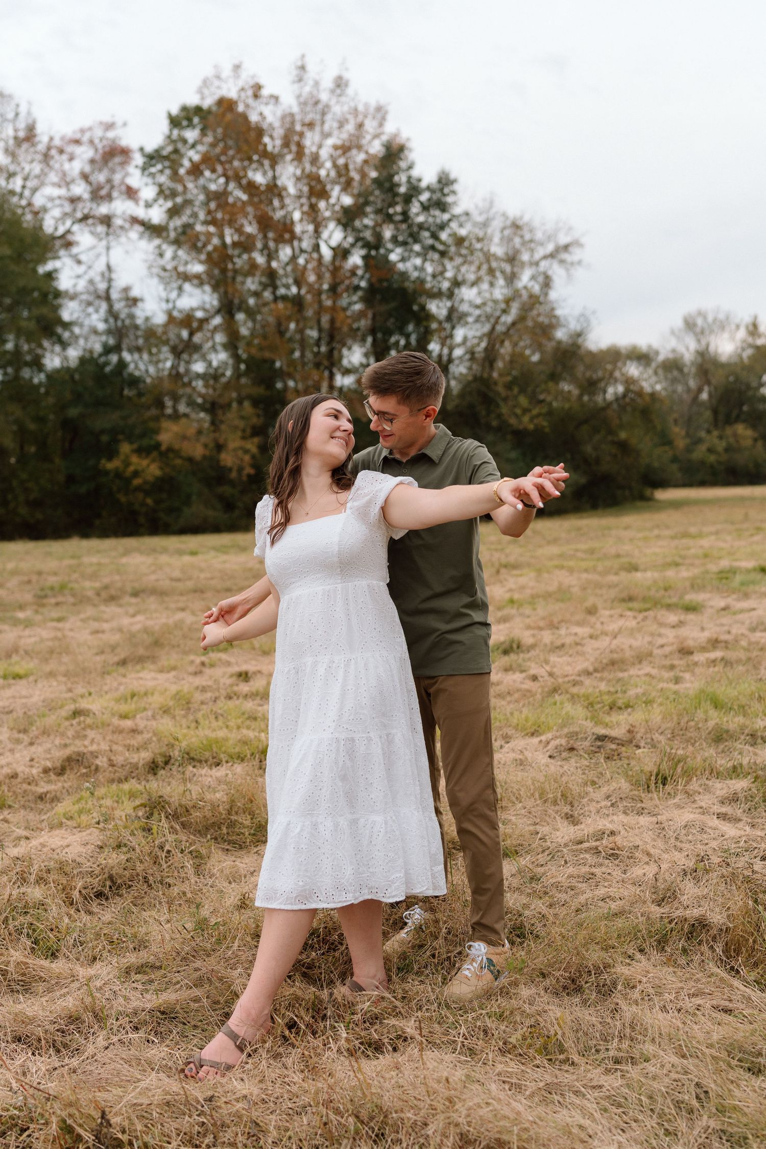 Couple dancing and laughing together in an open field with dry grass and autumn trees in background.