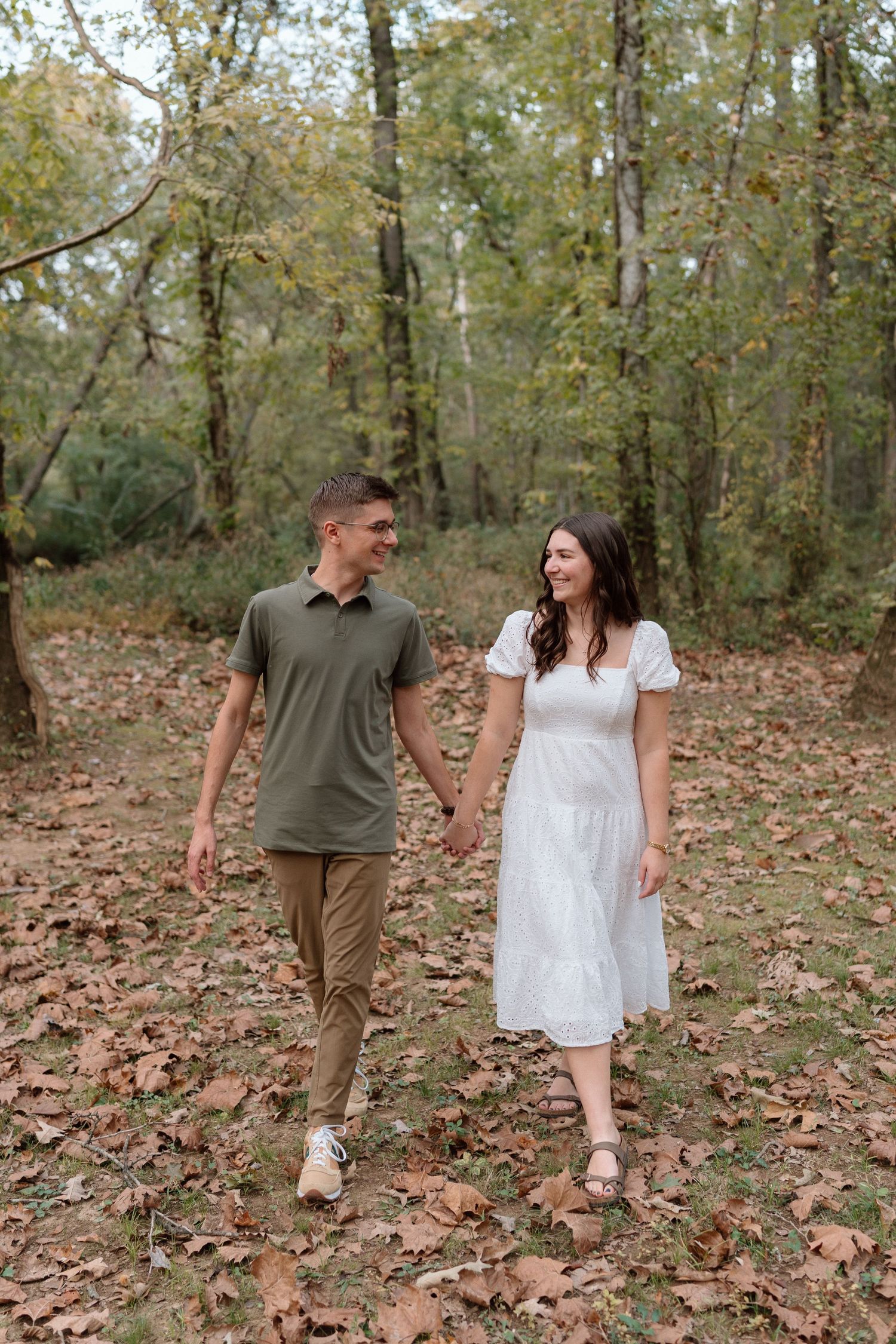 Couple holding hands walking through autumn forest with fallen leaves, man in green polo and woman in white dress.