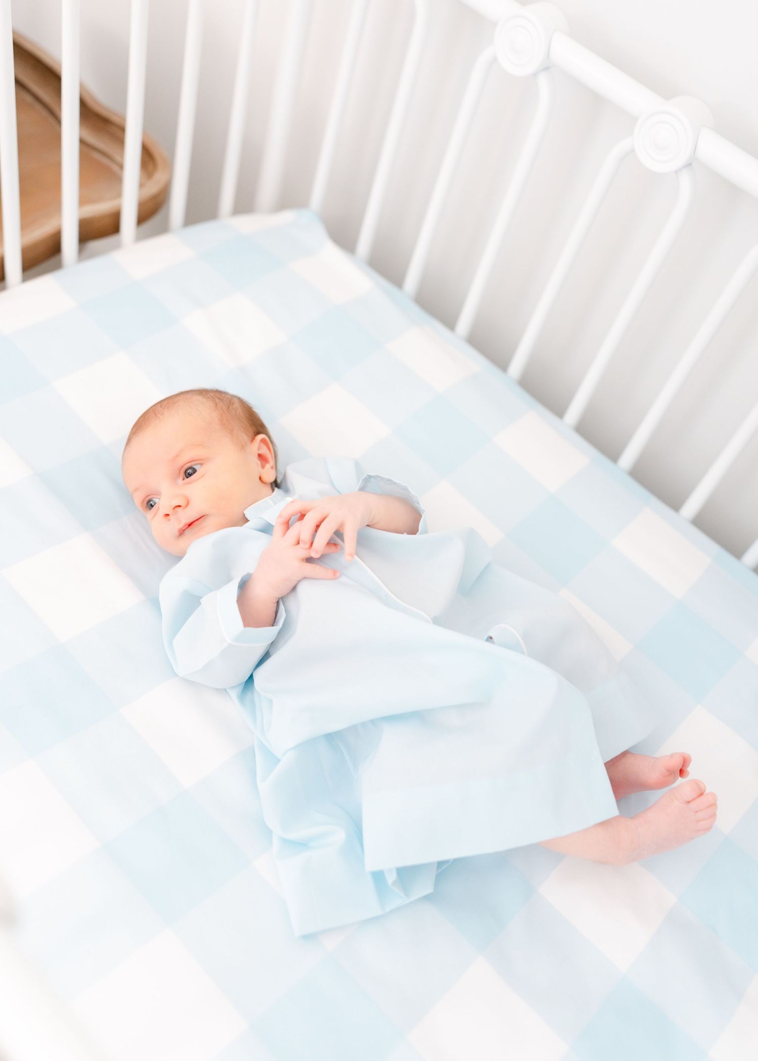 Newborn baby in blue sleeper lying on blue gingham crib sheet in white crib.