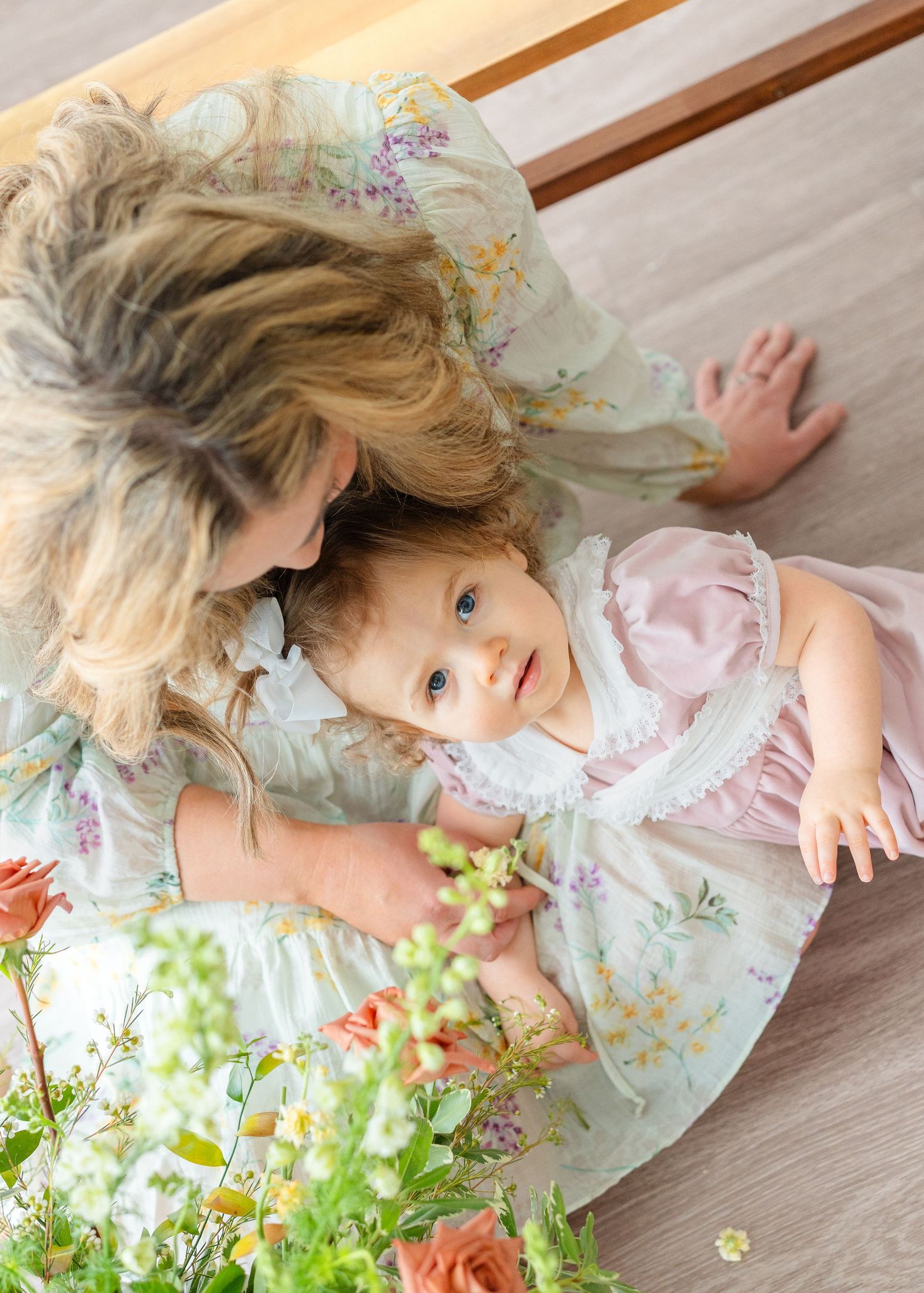 A mother in a floral dress leans over her baby girl lying on a wooden floor surrounded by flowers.