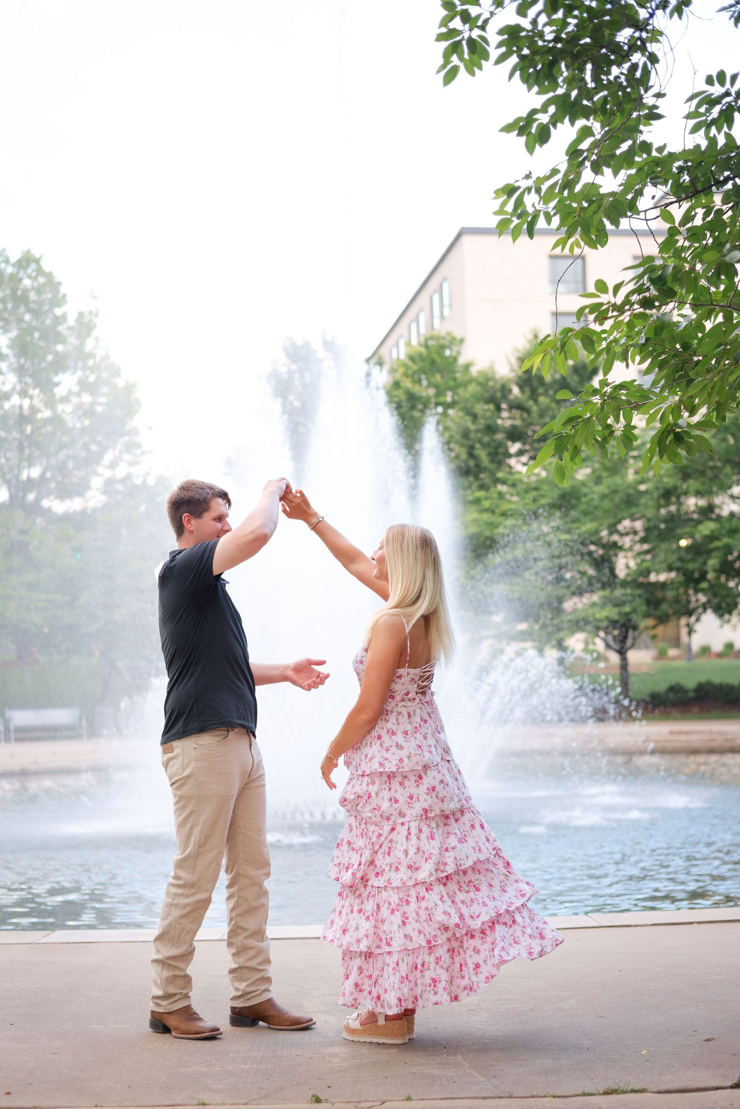 A couple dances near a fountain outdoors, the woman wearing a pink floral tiered maxi dress.