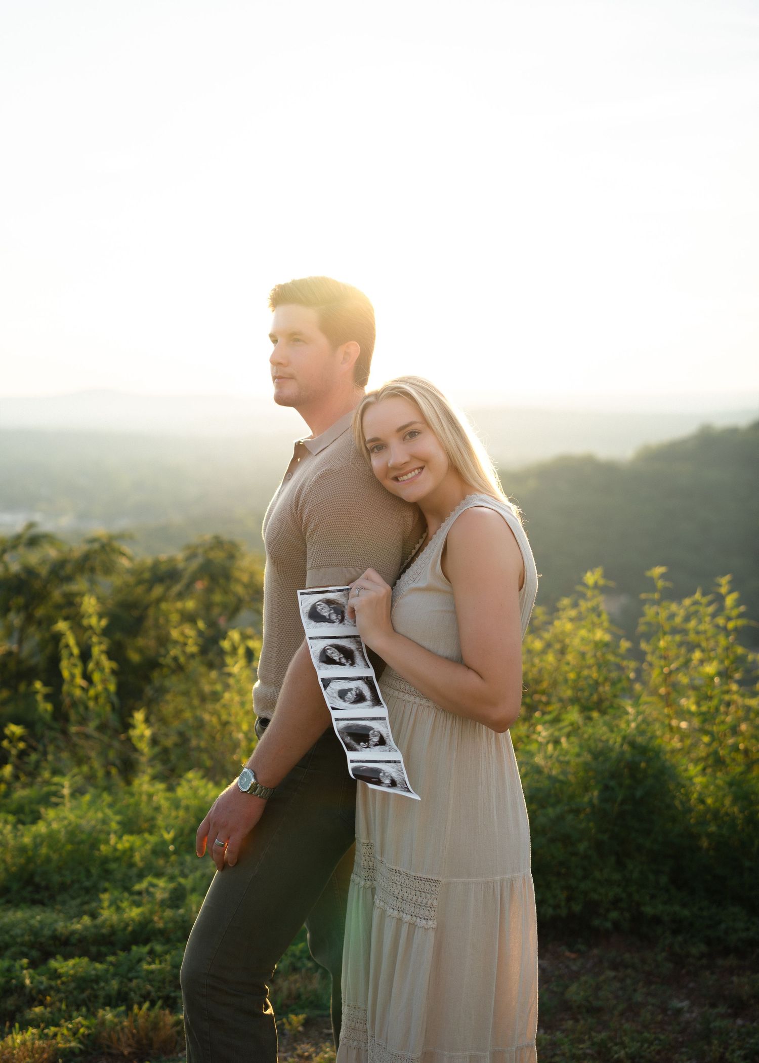 Couple holding ultrasound photos at golden hour overlook, pregnancy announcement portrait outdoors.