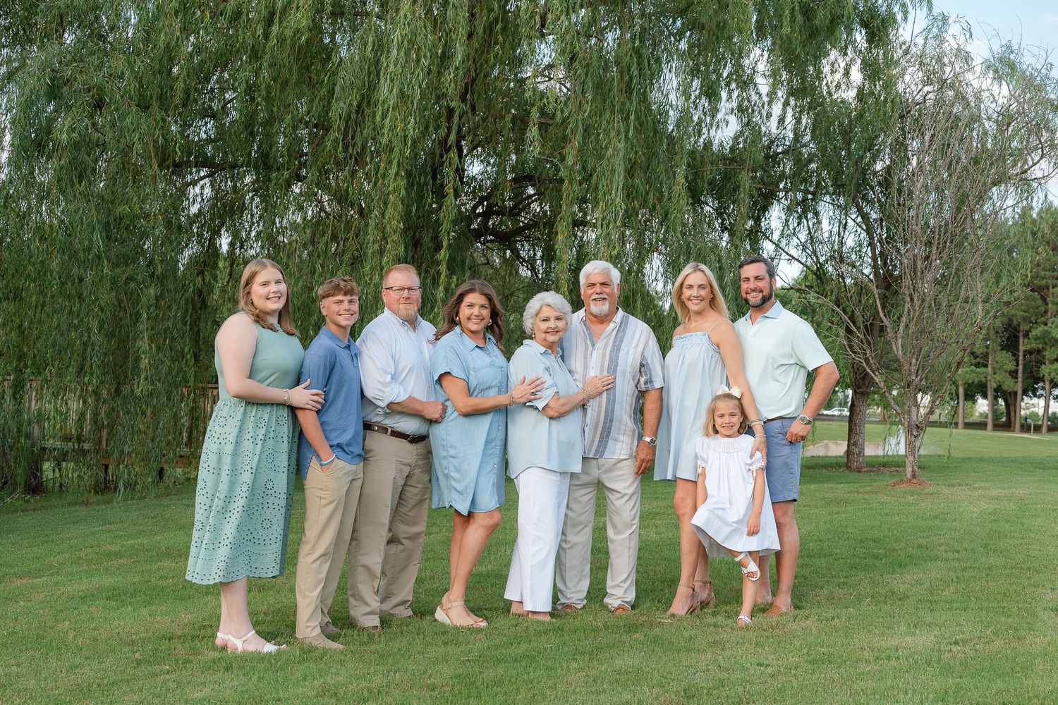 Family of eight poses outdoors in coordinated blue and green outfits under a willow tree on green grass.