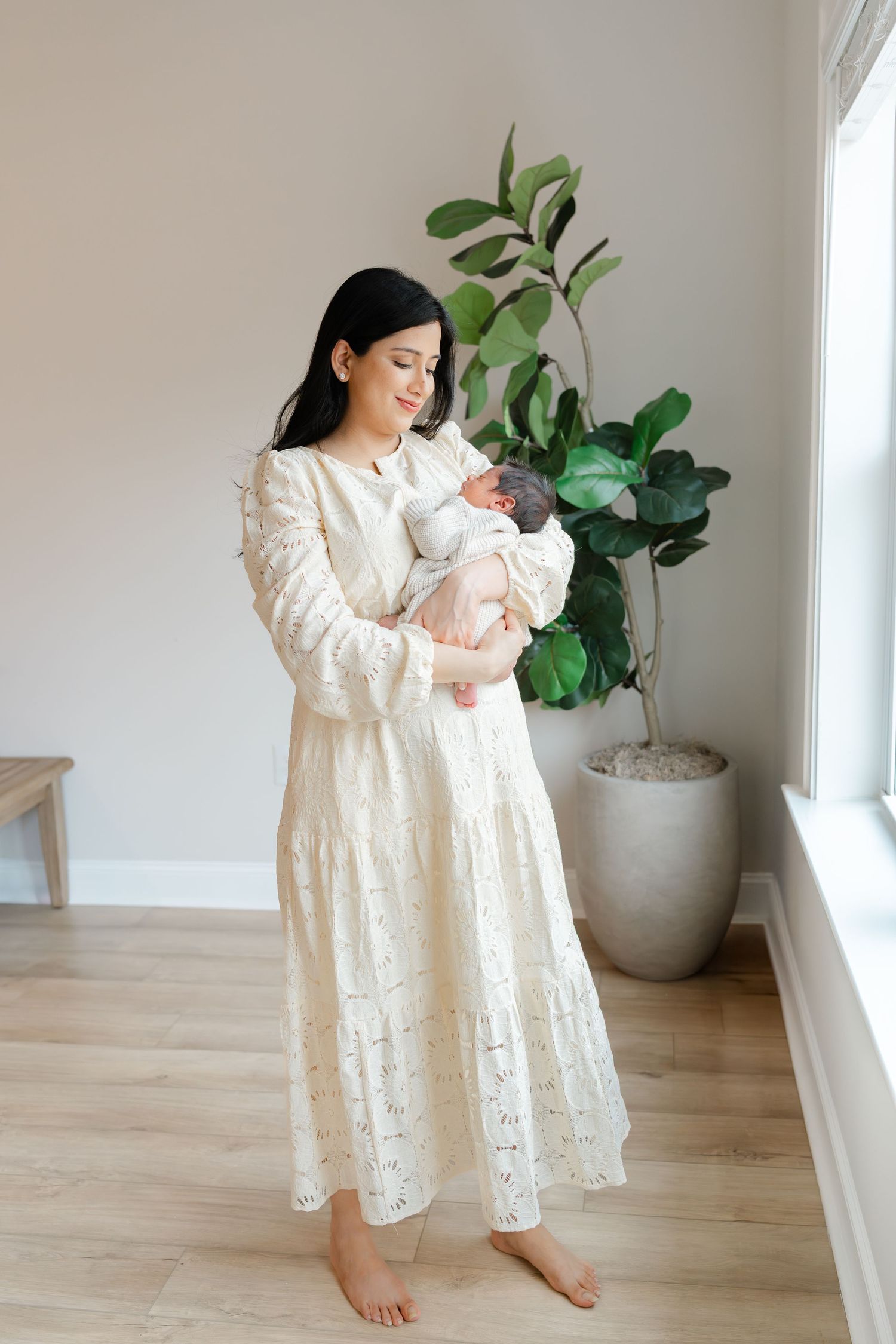 A dark-haired woman in a cream lace maxi dress holds her newborn baby while standing barefoot near a potted plant.