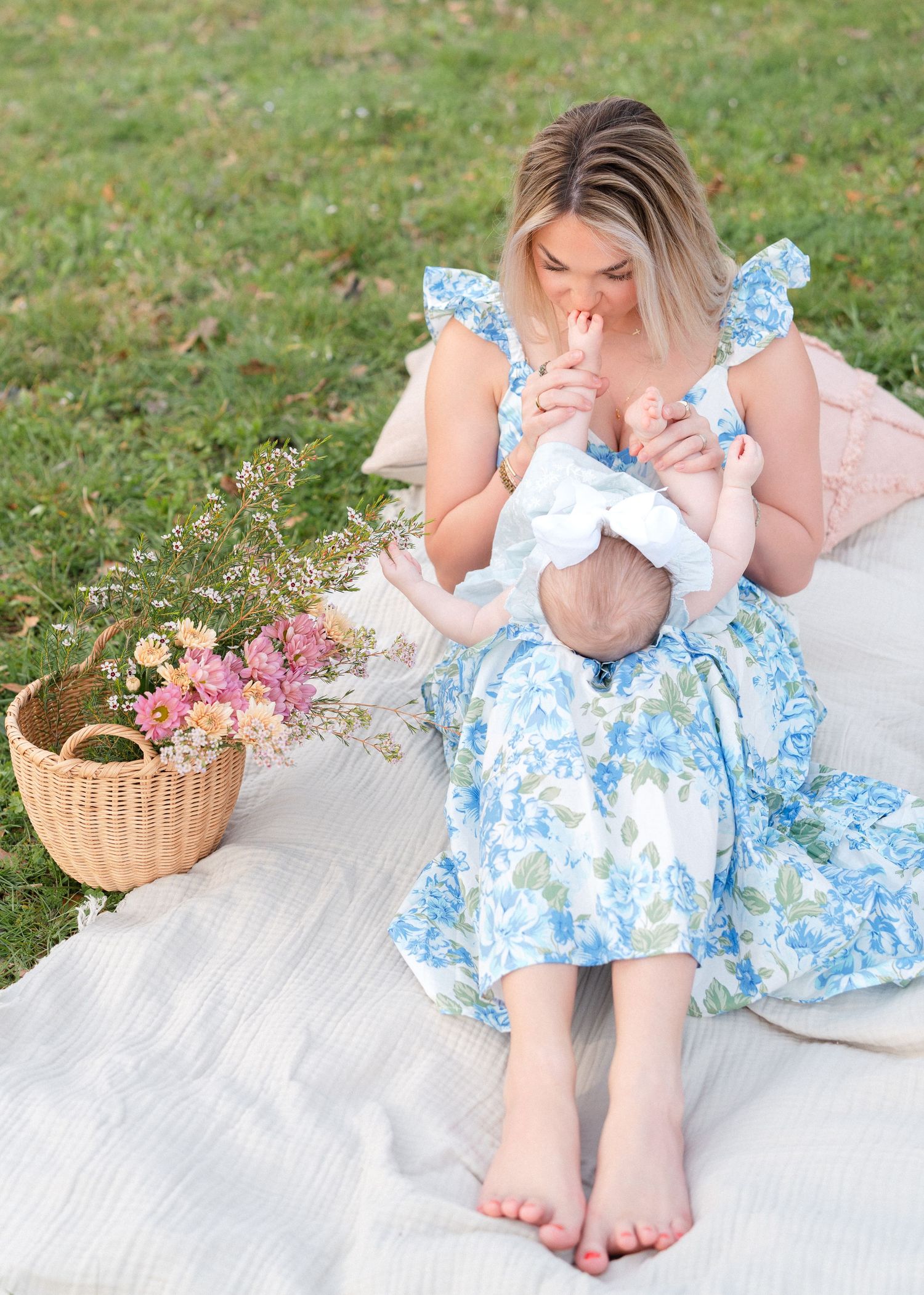 A barefoot mother in a blue floral dress kisses her baby's hand on an outdoor picnic blanket.