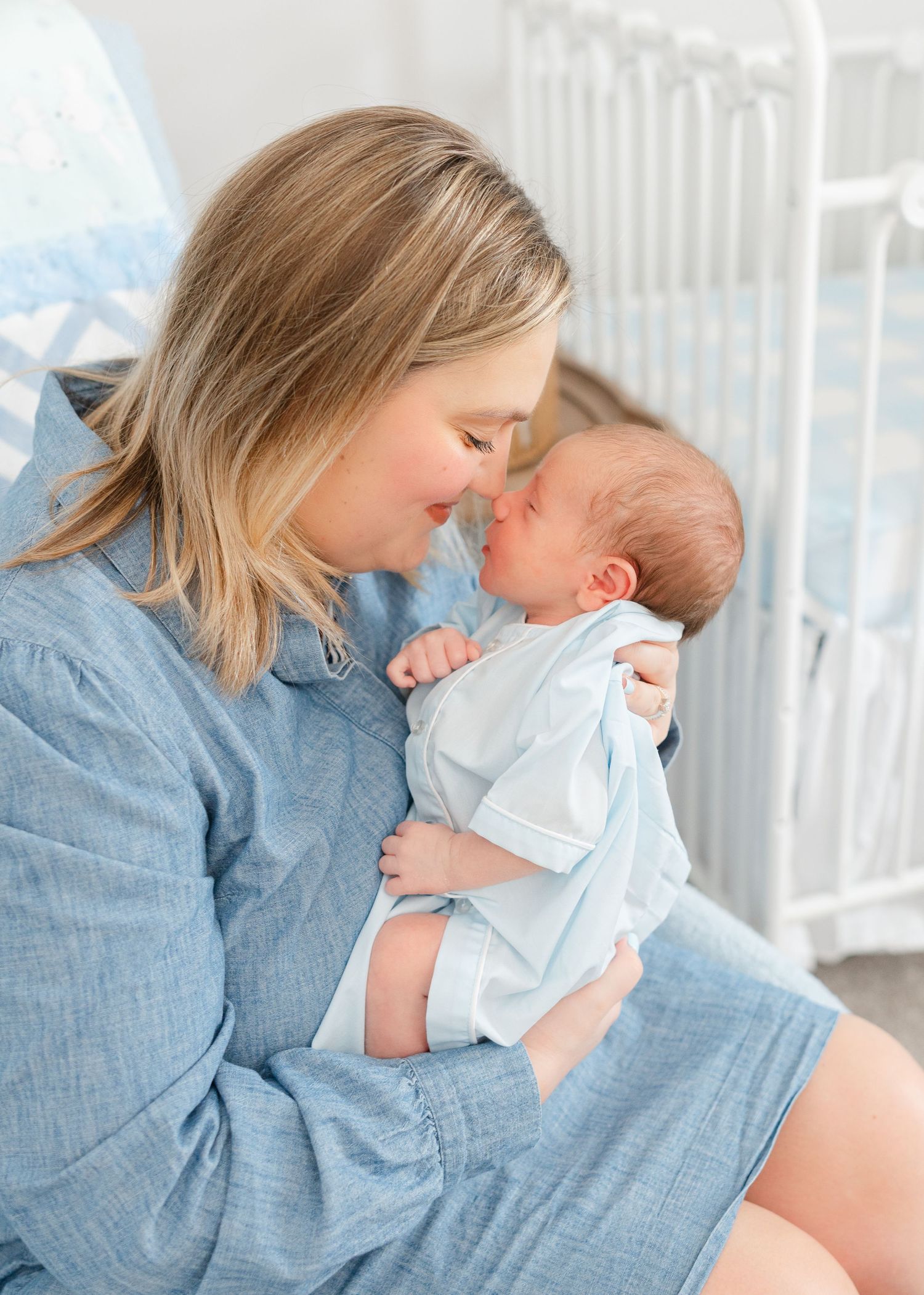 A mother tenderly touches noses with her newborn baby boy in a soft blue nursery setting.